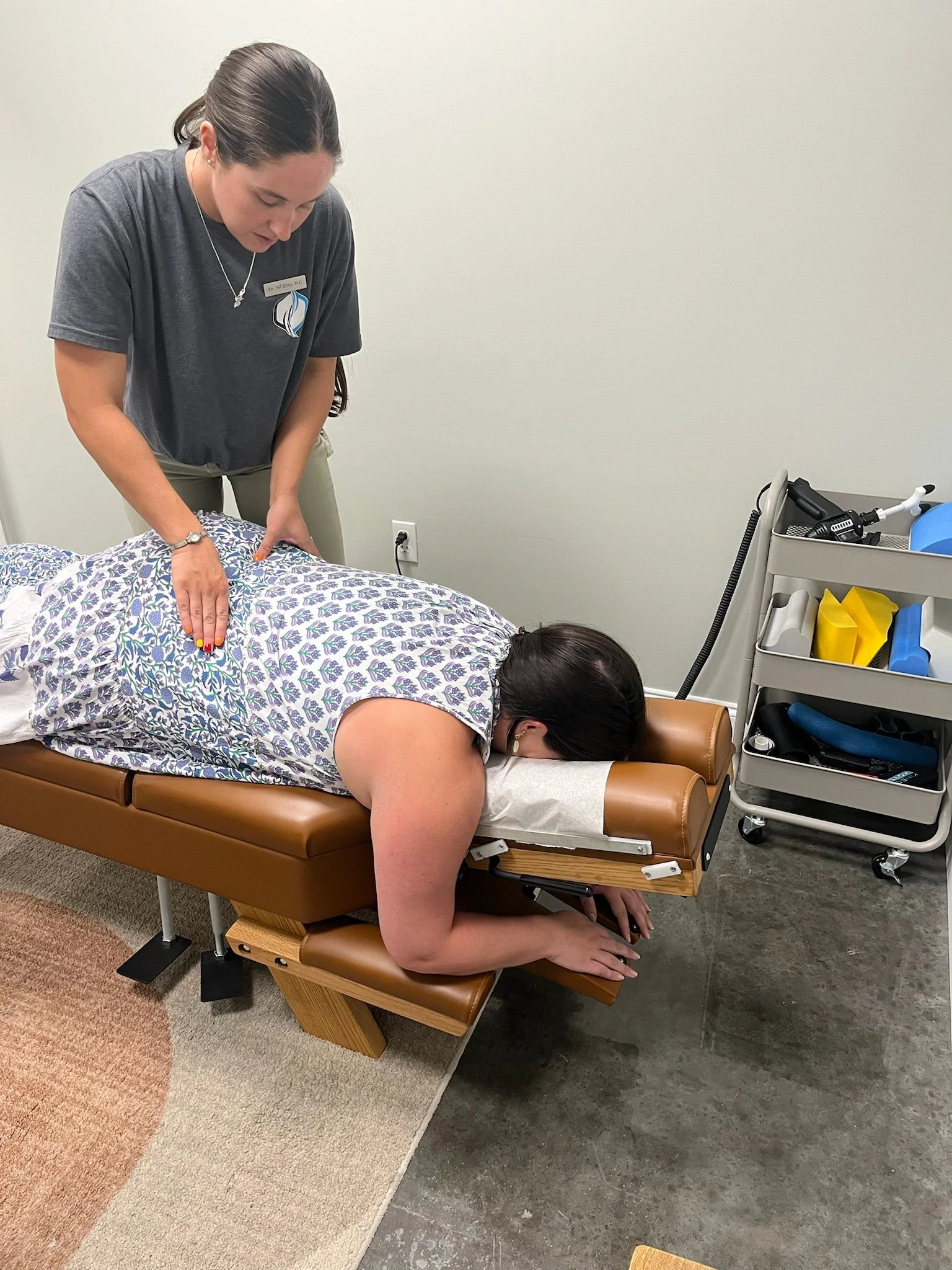 A woman is lying face down on a massage or chiropractic table, with her head turned to the side and her arm hanging off the side of the table. A healthcare professional is standing next to her, applying pressure to her back. The scene appears to be in a clinic or treatment room with medical equipment nearby.