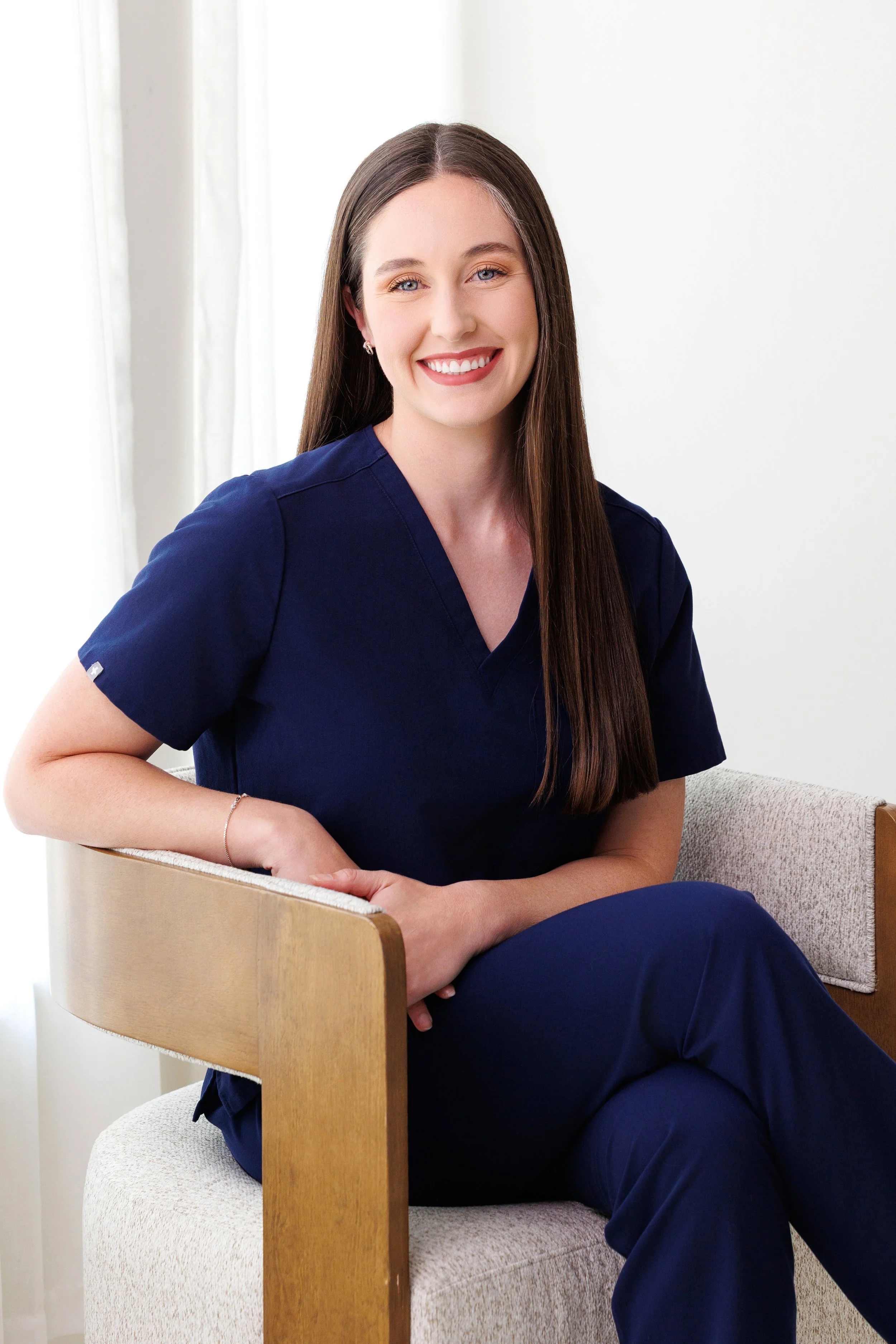 A woman with long brown hair, light skin, and blue eyes smiling, wearing navy medical scrubs, sitting on a beige chair with a wooden backrest, in a room with white walls and curtains.