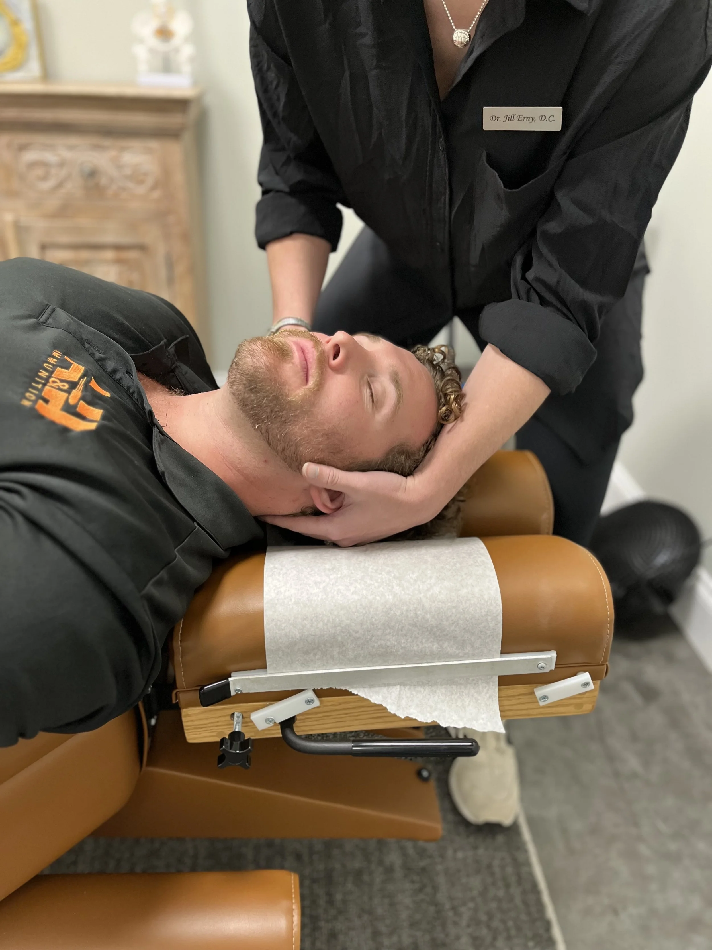 A person with a beard lying on a chiropractic adjustment table, receiving neck support treatment from a practitioner. The practitioner is wearing a black uniform with a name tag reading 'Dr. Jill Emry, D.C.' and a necklace. The setting appears to be a medical or chiropractic office.