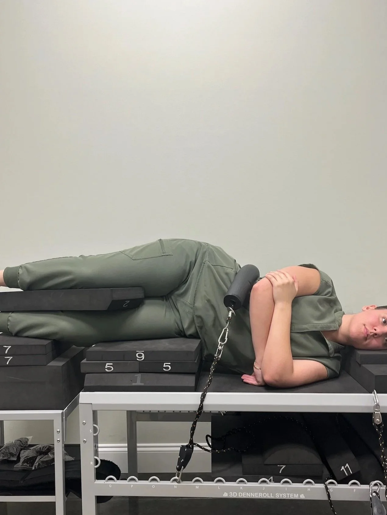 A person lying on a training table in a physical therapy or rehabilitation setting, with their arms crossed over their chest, their head turned to the side, and a chain attached to their ankle.