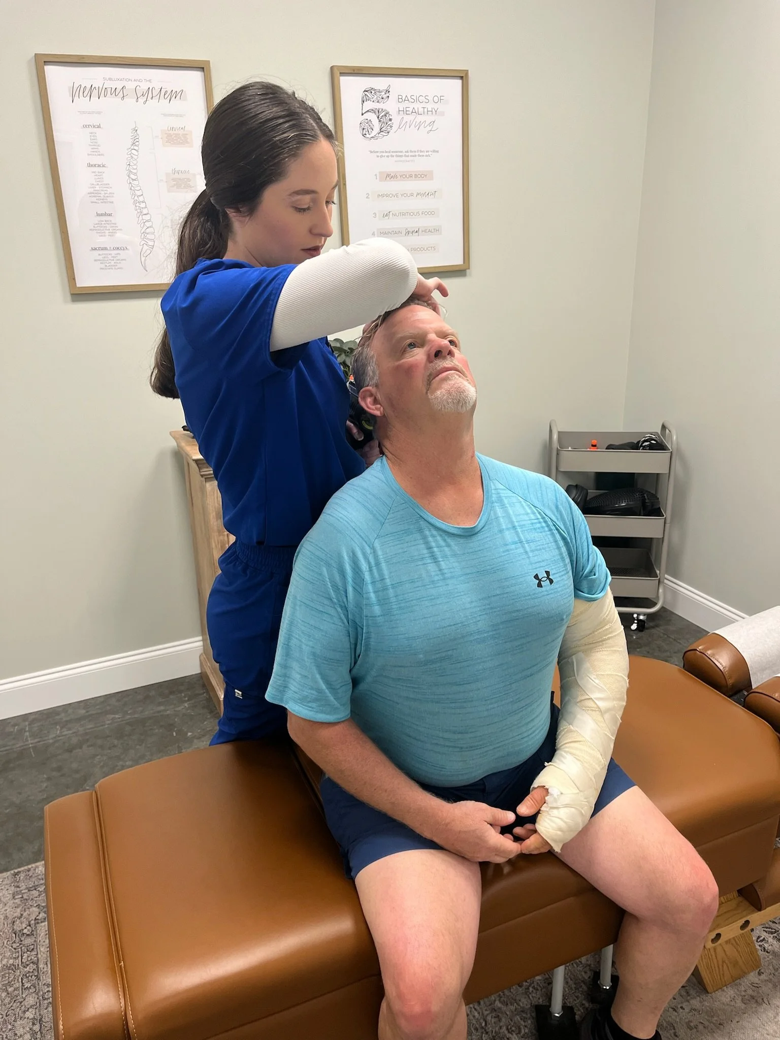 A woman with long brown hair wearing a blue medical uniform is examining a man, who has a cast on his arm and a sleeve over his other arm. The man is sitting on a brown examination table in a medical office, looking upward. There are framed educational posters on the wall behind them.