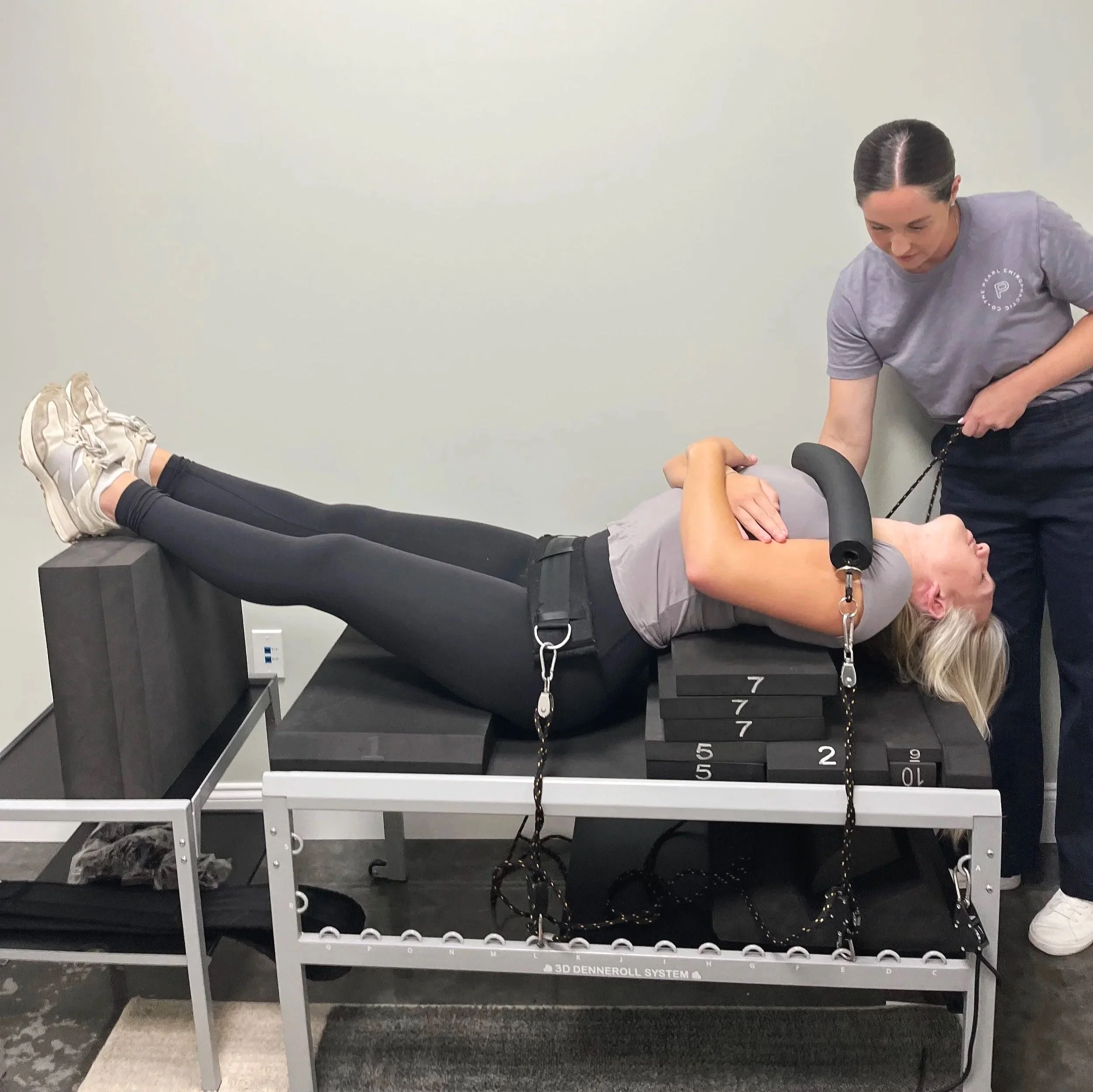 A woman providing physical therapy to a person lying on a treatment table, with resistance bands attached to their arm, in a clinical setting.