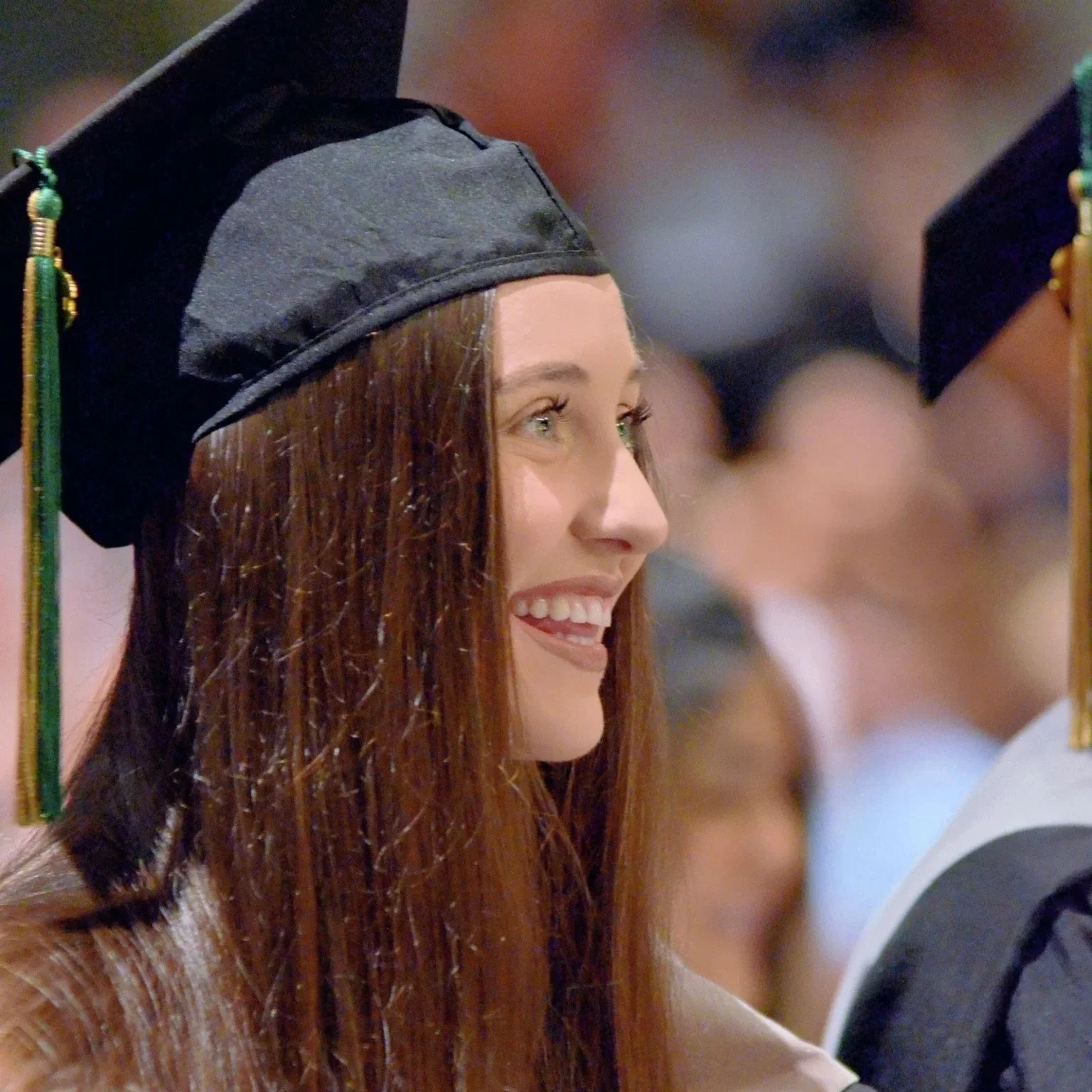 A young woman in a black graduation cap and gown is smiling during a graduation ceremony.