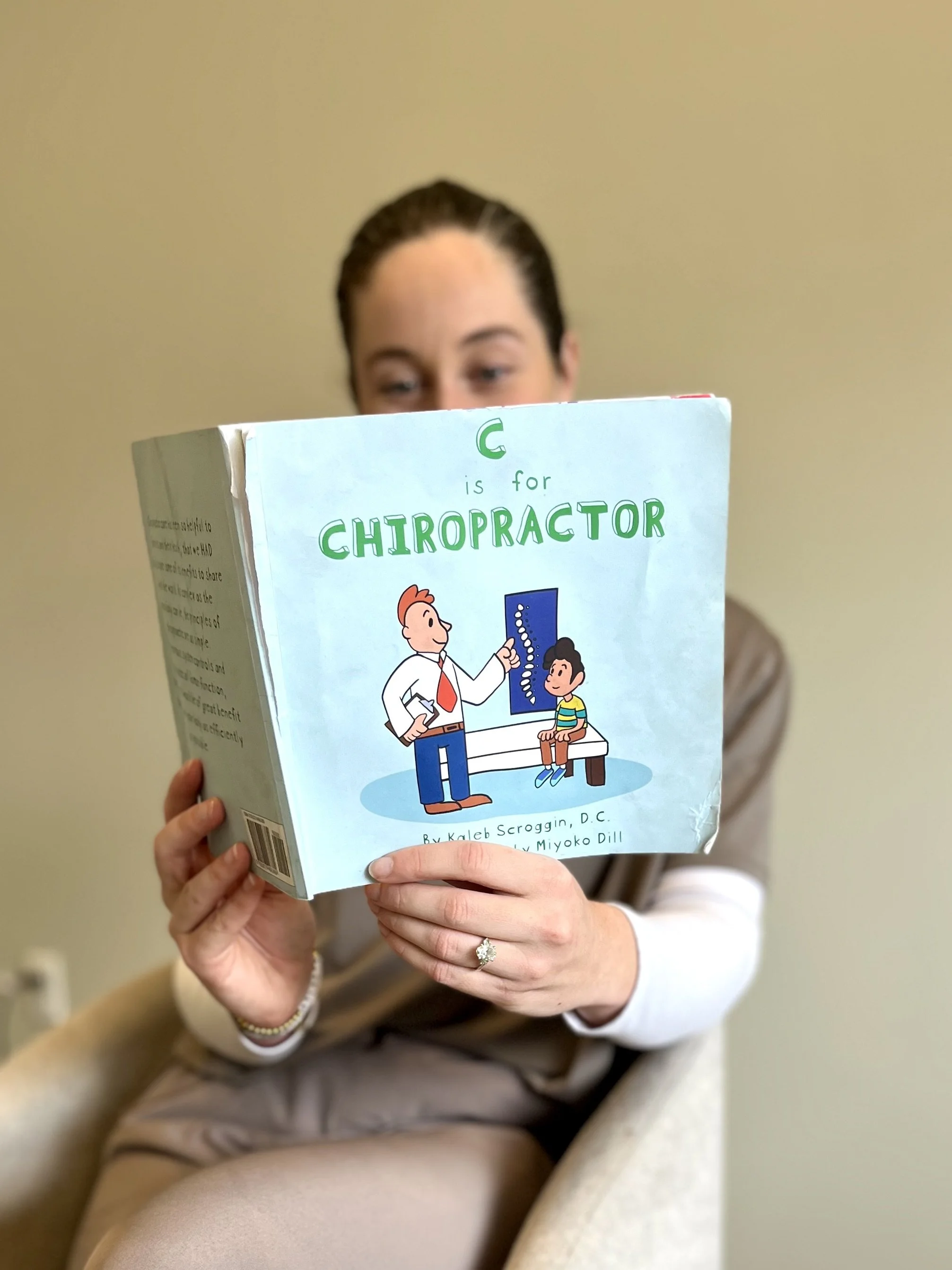 A woman with brown hair holding a children's book titled 'C is for Chiropractor' while sitting in a beige armchair. The book features an illustration of a chiropractor adjusting a child's spine.