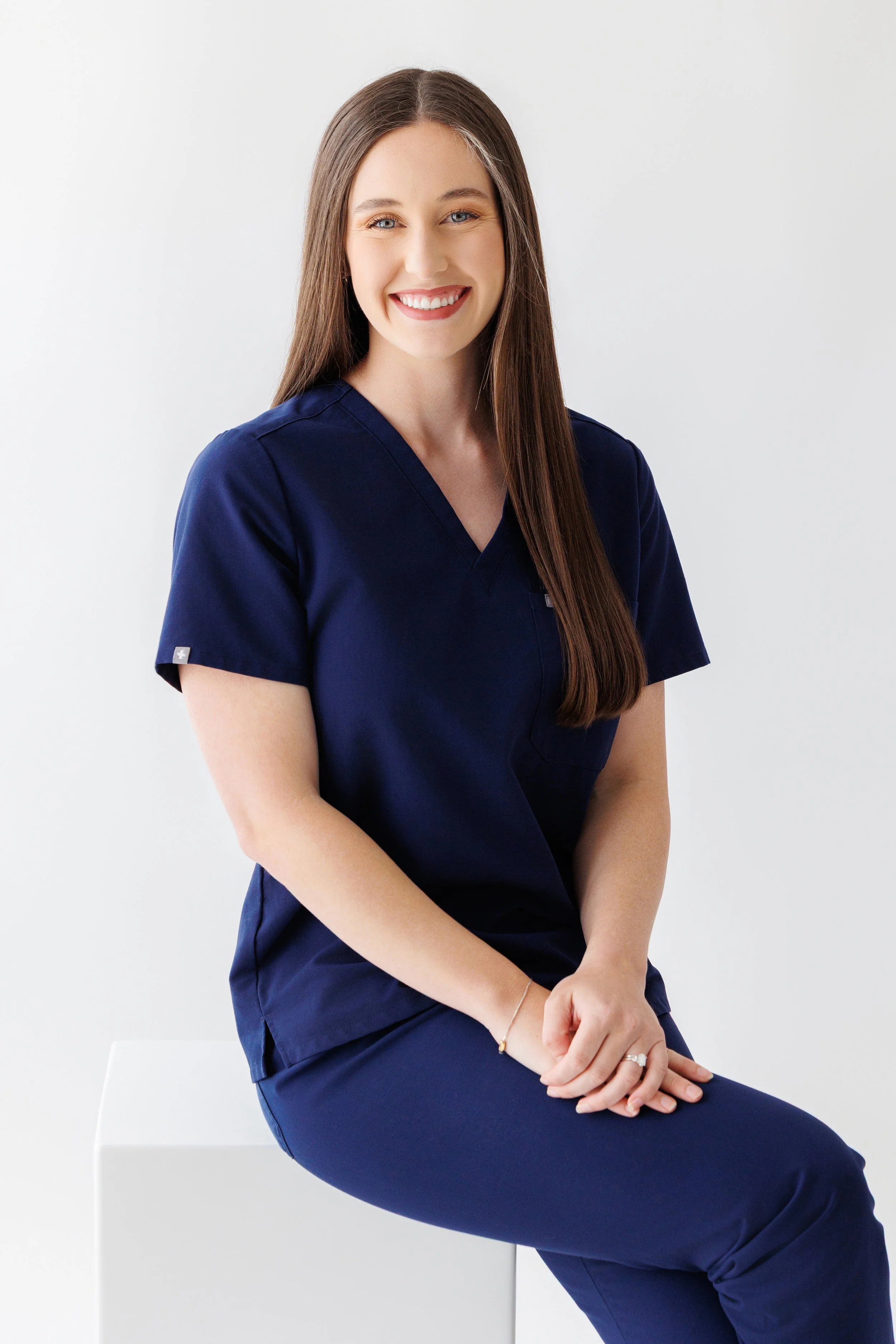 A woman with long brown hair smiling, wearing navy scrubs, sitting on a white stool with her hands clasped, against a plain white background.