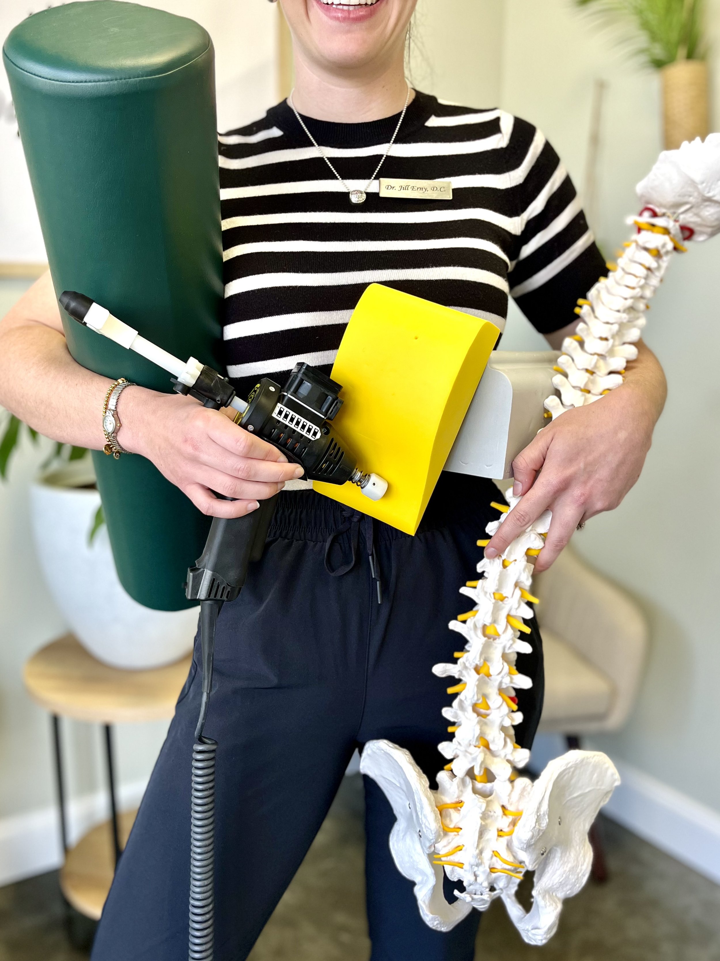 A healthcare professional holding a model of a human spine and pelvis while standing in a clinic or office setting.