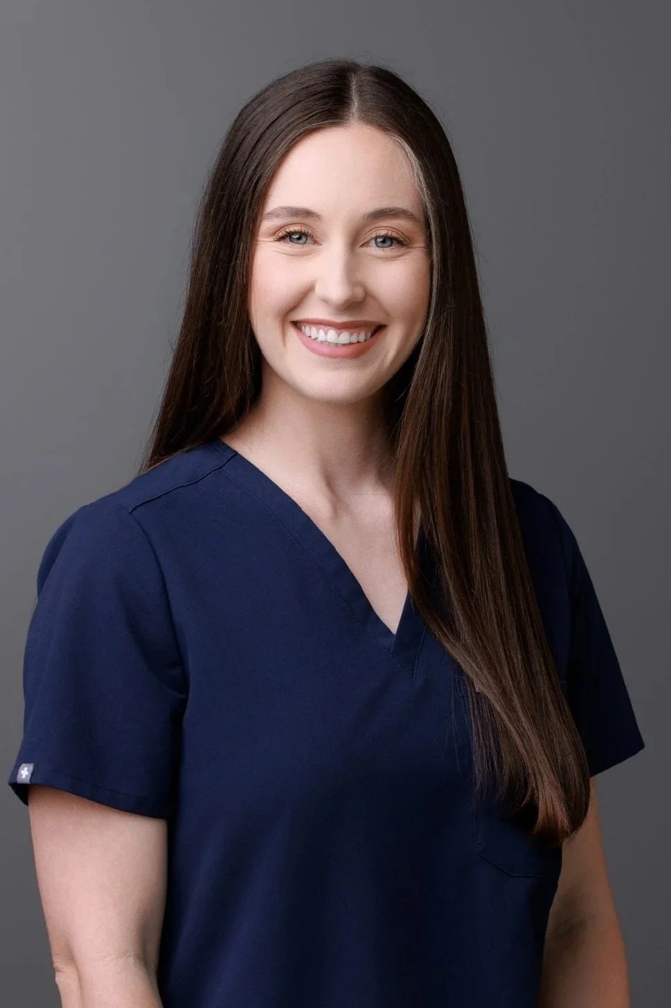 A young woman with long brown hair, smiling, wearing navy scrubs, against a gray background.