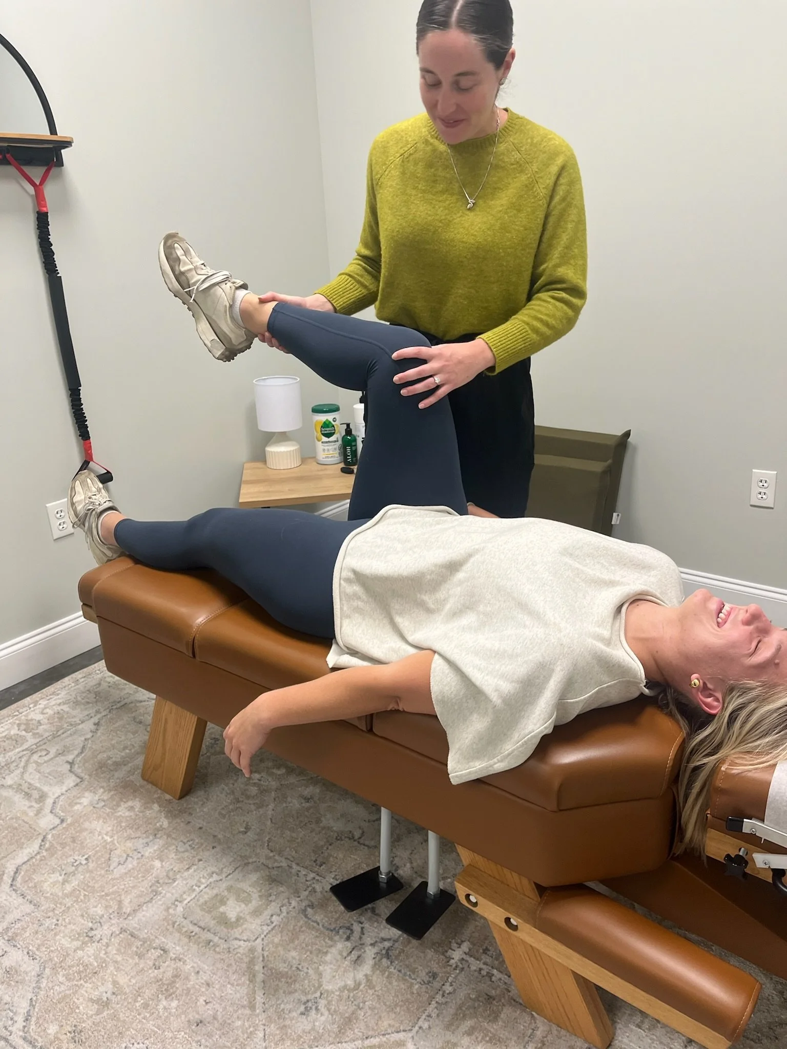 A woman lying face down on a massage table with her eyes closed, smiling. A therapist is holding one of her legs, likely performing a physical therapy or massage procedure. The setting appears to be a clinic or therapy room with a neutral decor and a small table with bottles and a lamp in the background.
