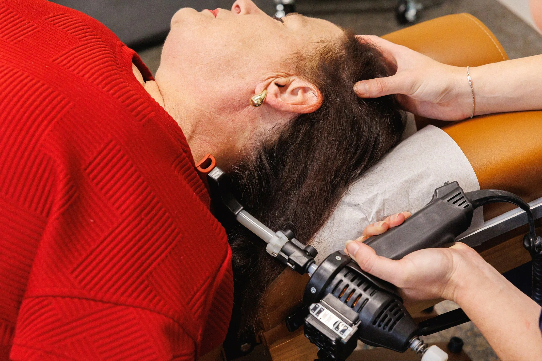 A woman lying on a massage table receives a chiropractic adjustment using a pneumatic tool. She is wearing a red sweater and has a gold earring, with someone applying the tool to her neck area.