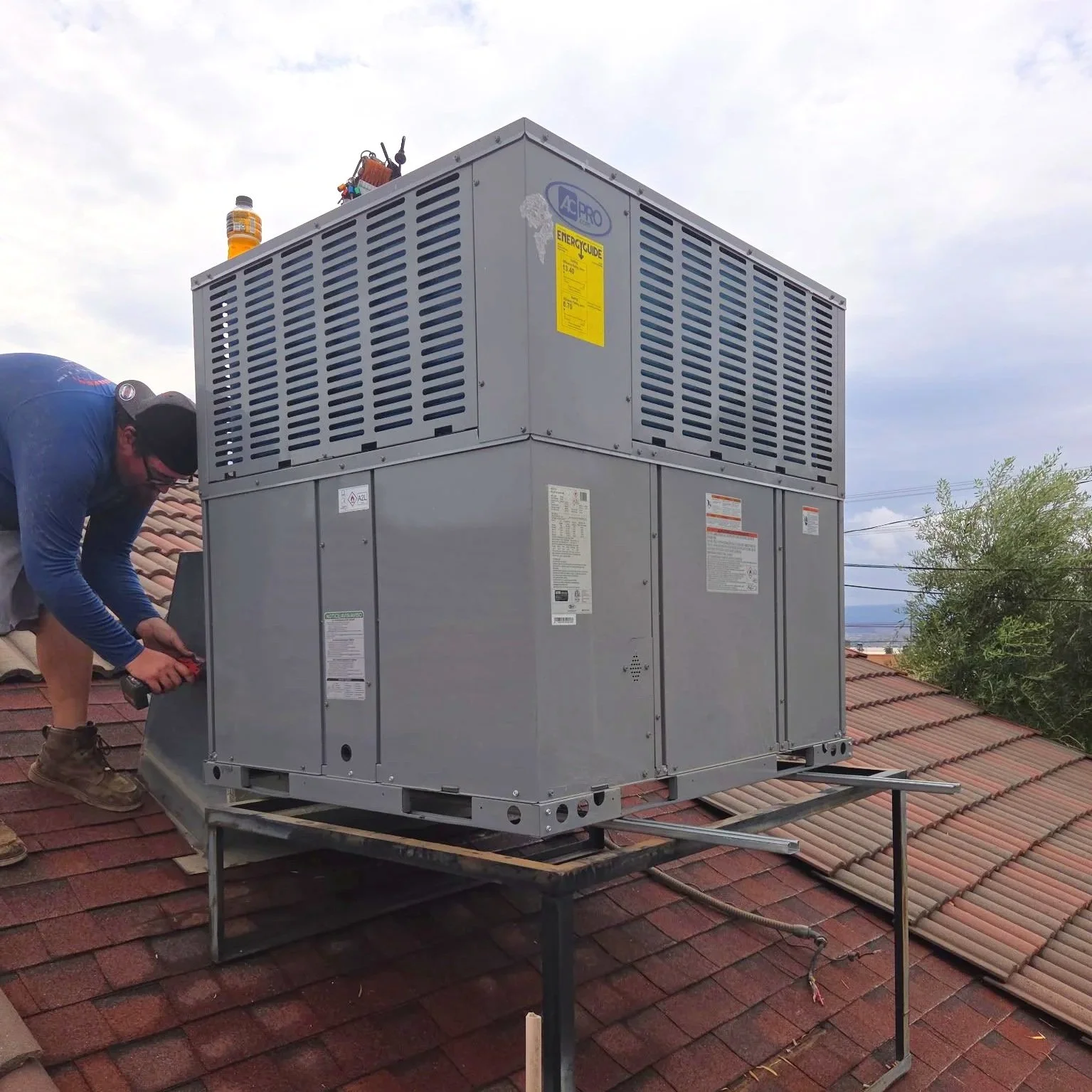A worker installing an HVAC unit on a residential rooftop with a person holding tools beside the unit and a bottle of orange drink on top of the unit.