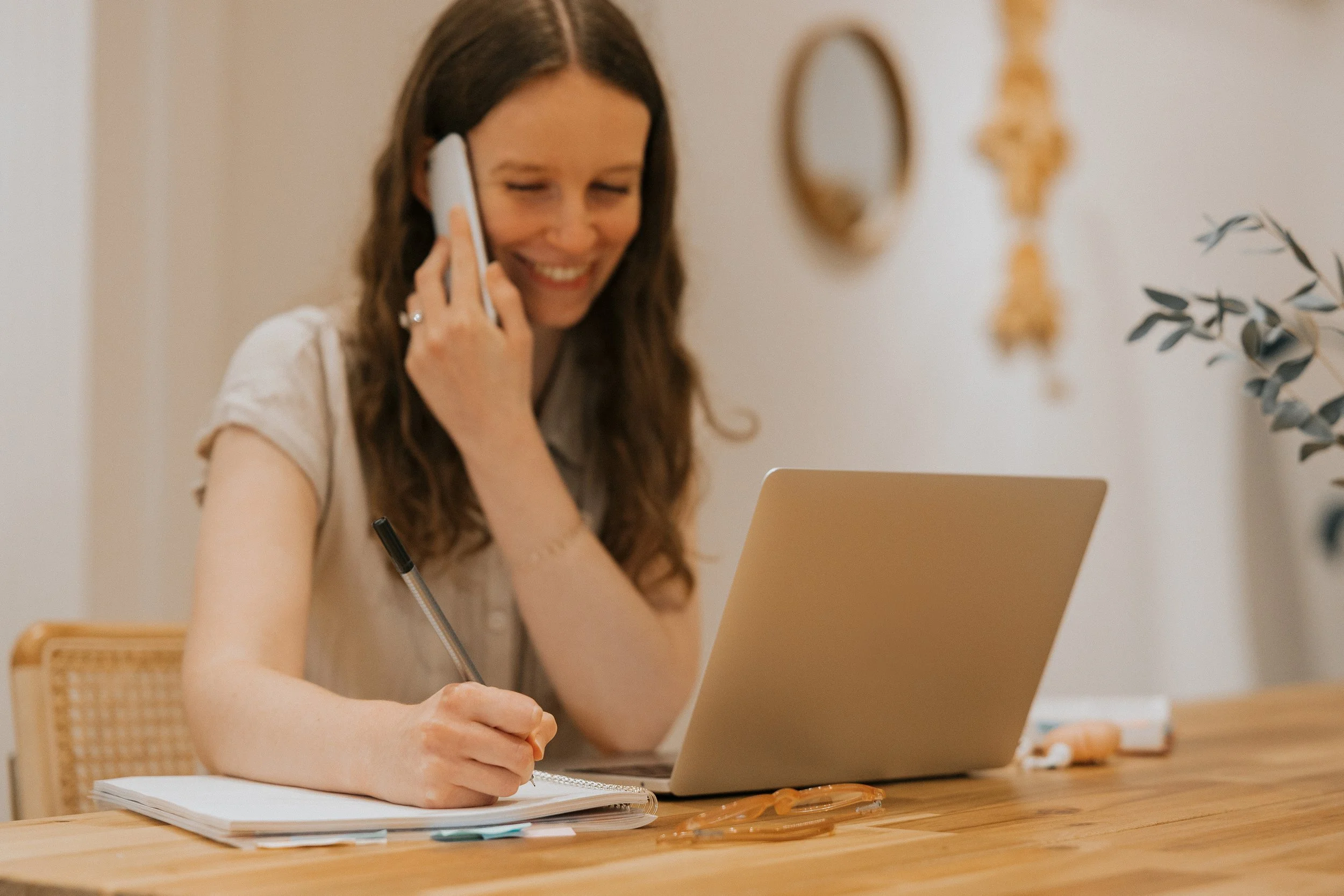 Woman on the phone with a client, taking notes on a notebook with her laptop open in front of her