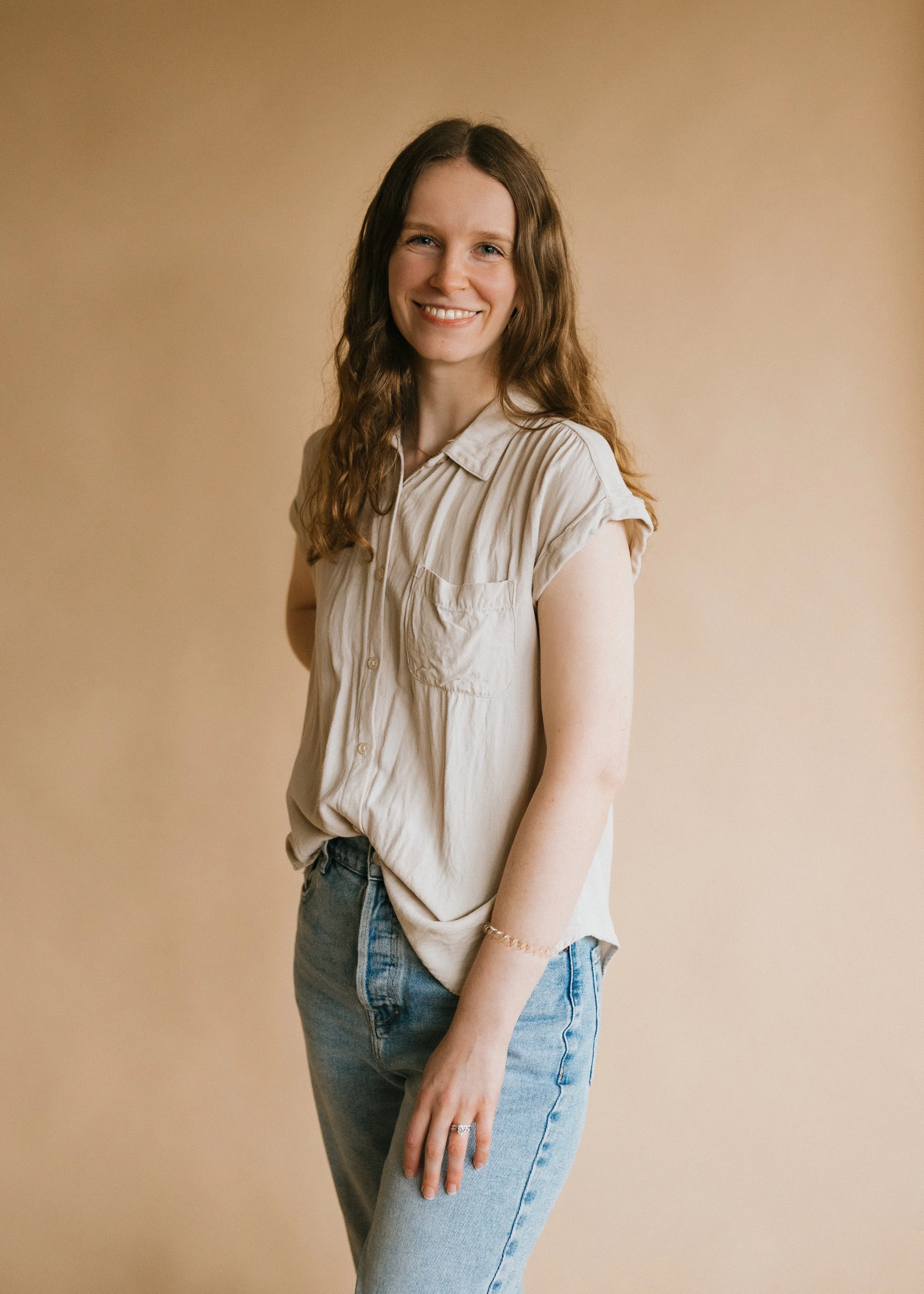 Photo of woman standing against a tan background with her hand on her hip, smiling at the camera