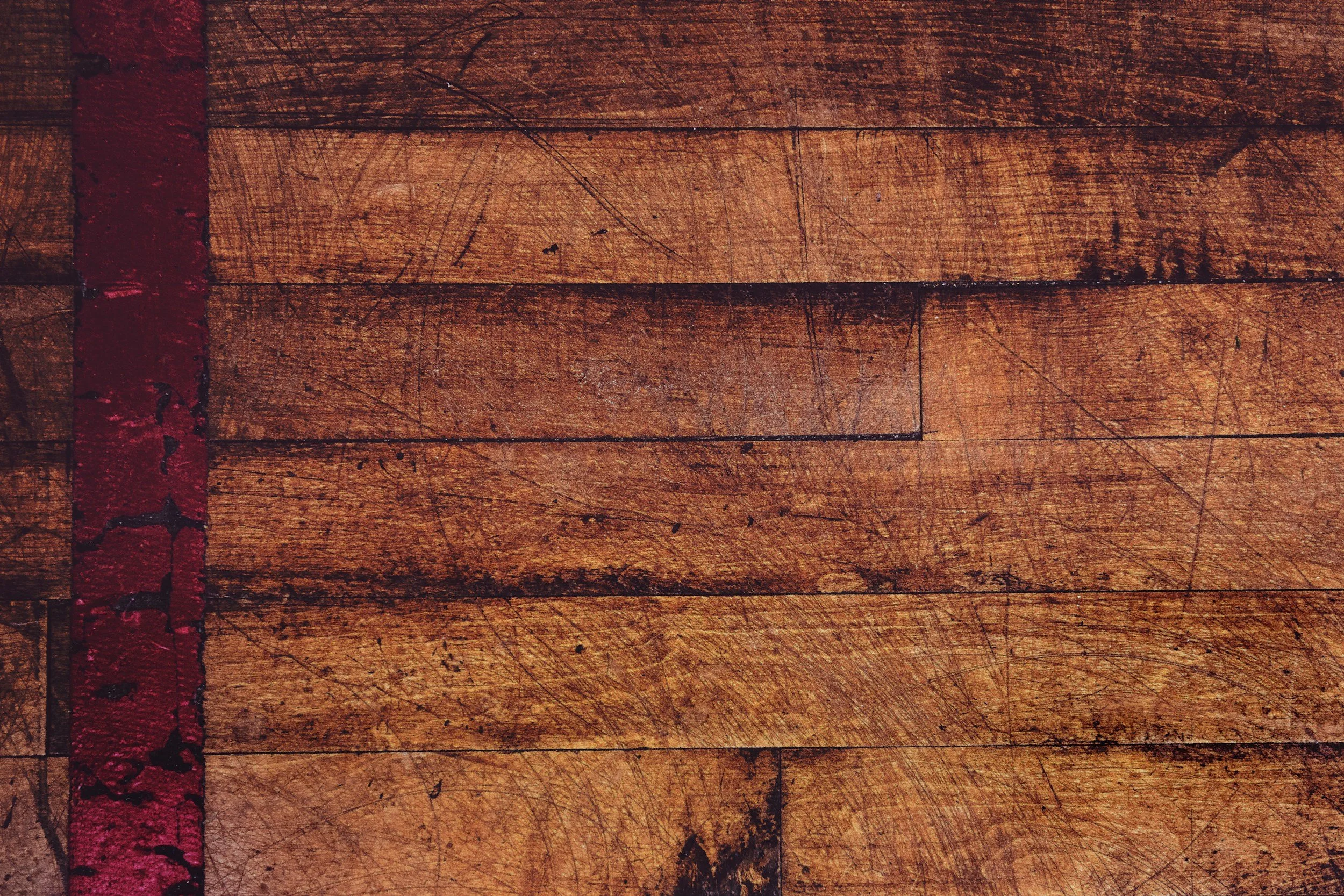 Close-up of wooden flooring with a red painted line on the left side, showing scratches and wear.