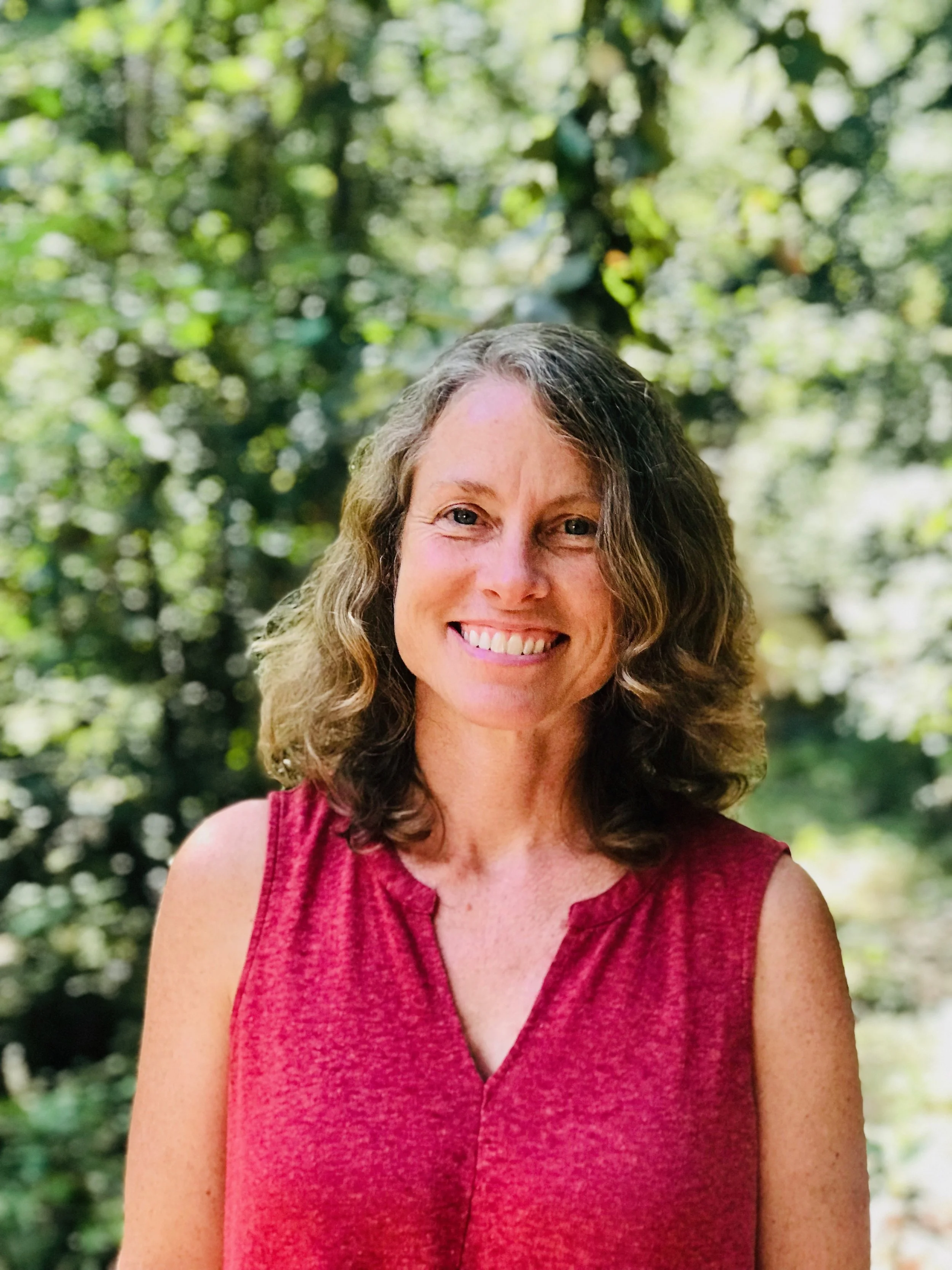 A woman with shoulder-length wavy hair smiling outdoors, wearing a sleeveless red top, with greenery in the background.