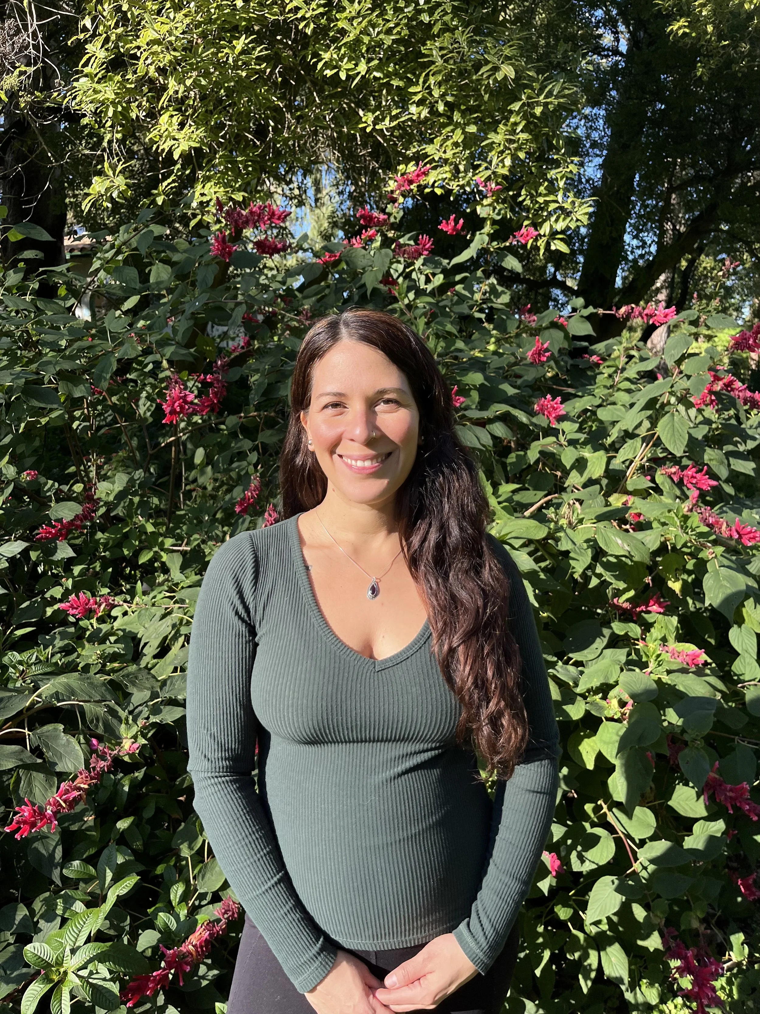 A woman with long brown hair smiling in front of a lush green bush with pink flowers on a sunny day.