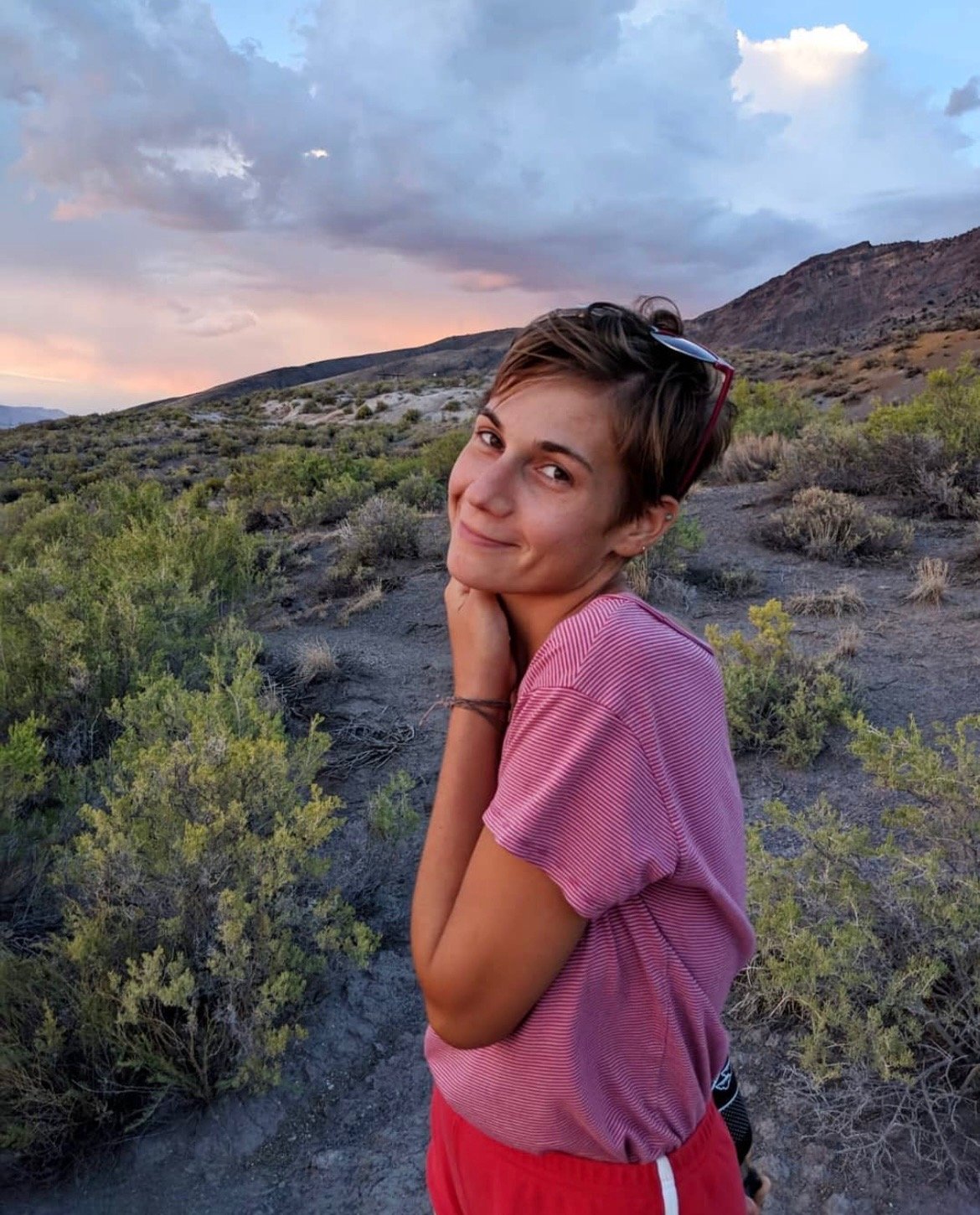 Young woman with short hair and sunglasses on her head smiling on a desert trail at sunset amidst dry shrubs and mountains.