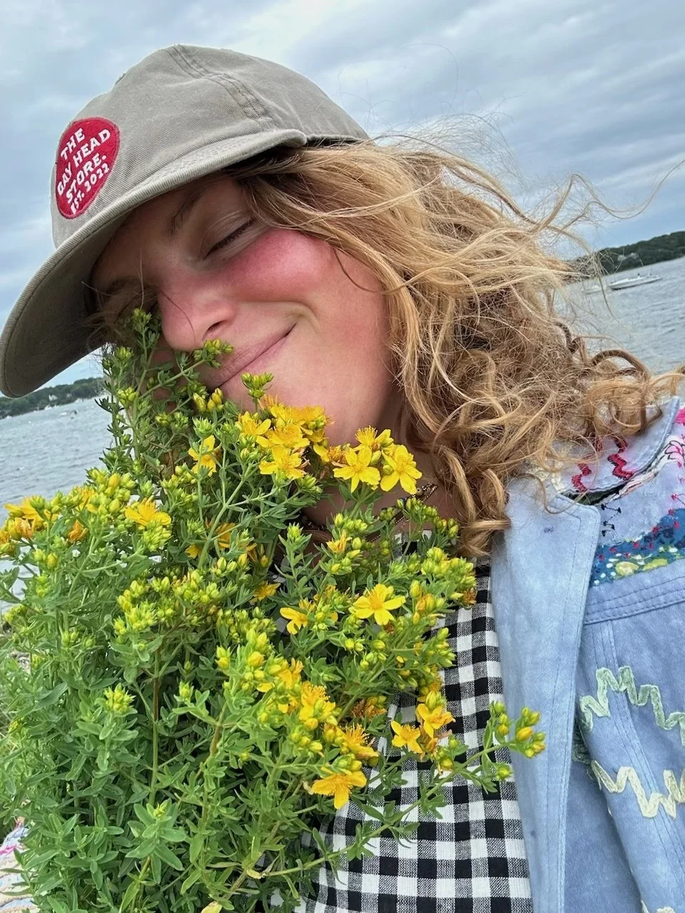 A woman with curly blonde hair wearing a gray hat and a denim jacket is smiling with her eyes closed and holding a large bouquet of yellow flowers near her face. She is outdoors near water with boats in the background.