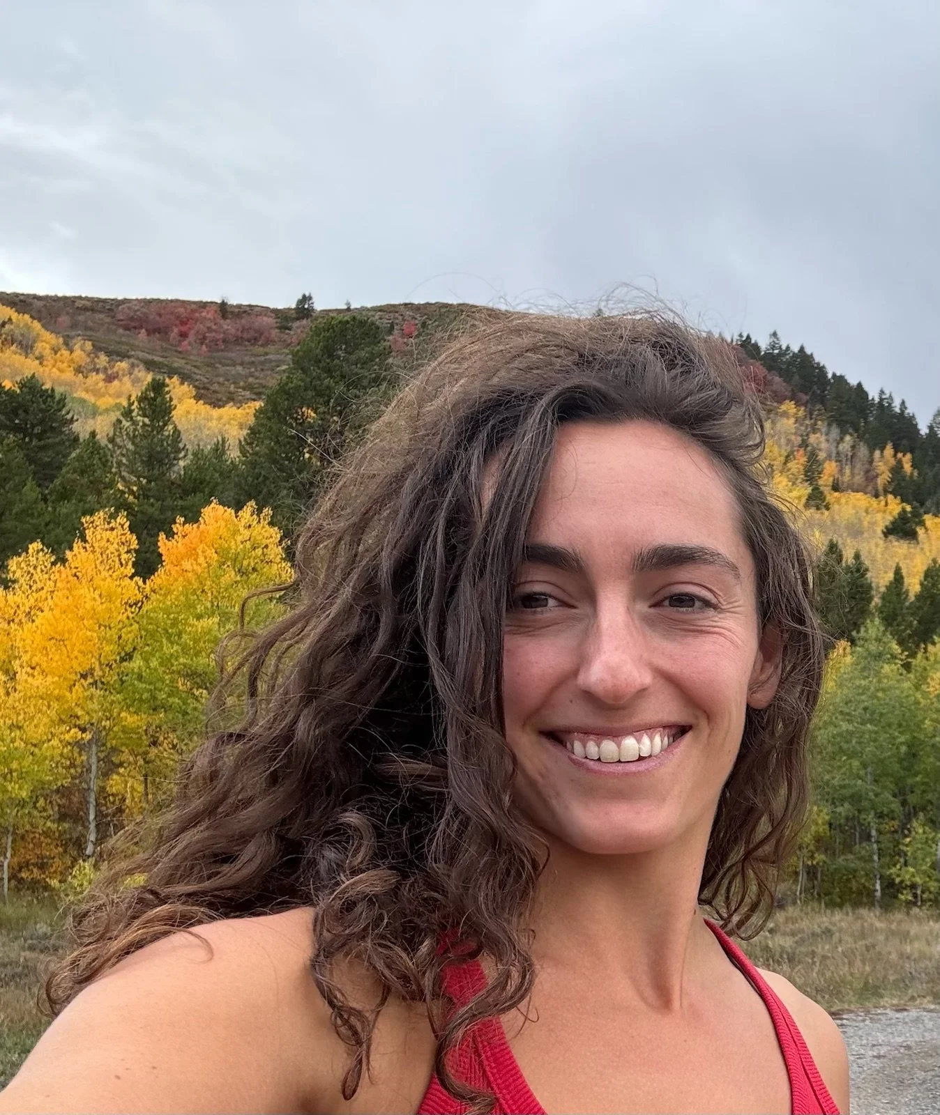 A smiling woman with curly brown hair wearing a red tank top outdoors with a background of trees with yellow and green leaves and a mountain under a cloudy sky.