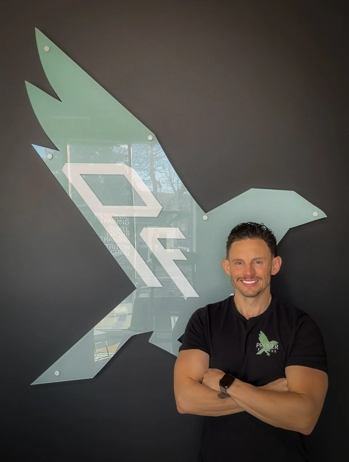 Man with crossed arms standing in front of a wall with a logo that looks like a stylized eagle and the words 'Proper Fitness' on his shirt.