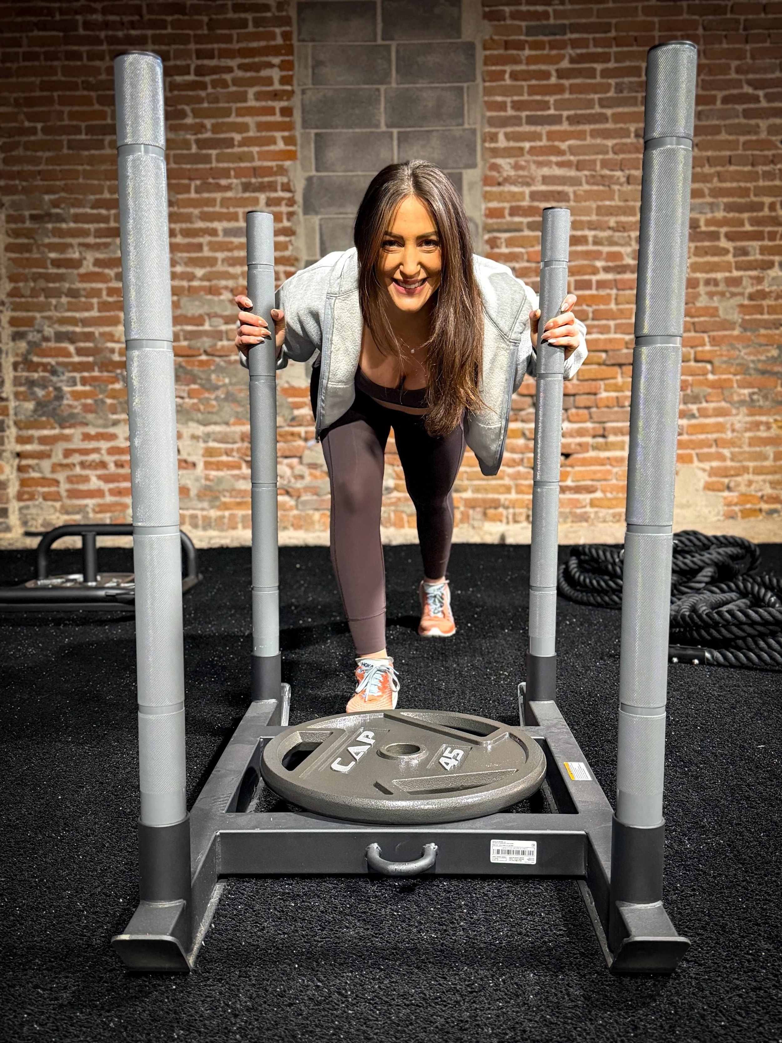 Woman smiling and exercising with a sled at a personal training gym with brick walls and black flooring.