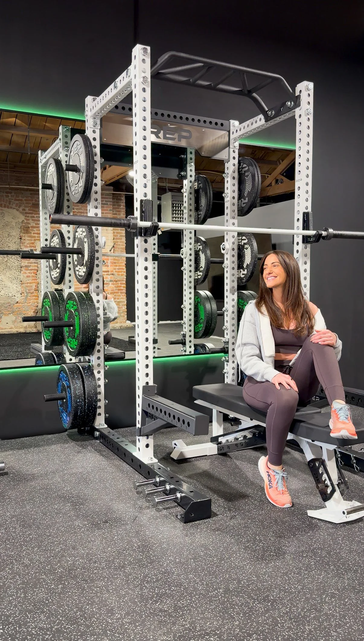 A woman sitting on a workout bench at the fitness studio with weightlifting equipment around her. Happy to see results.