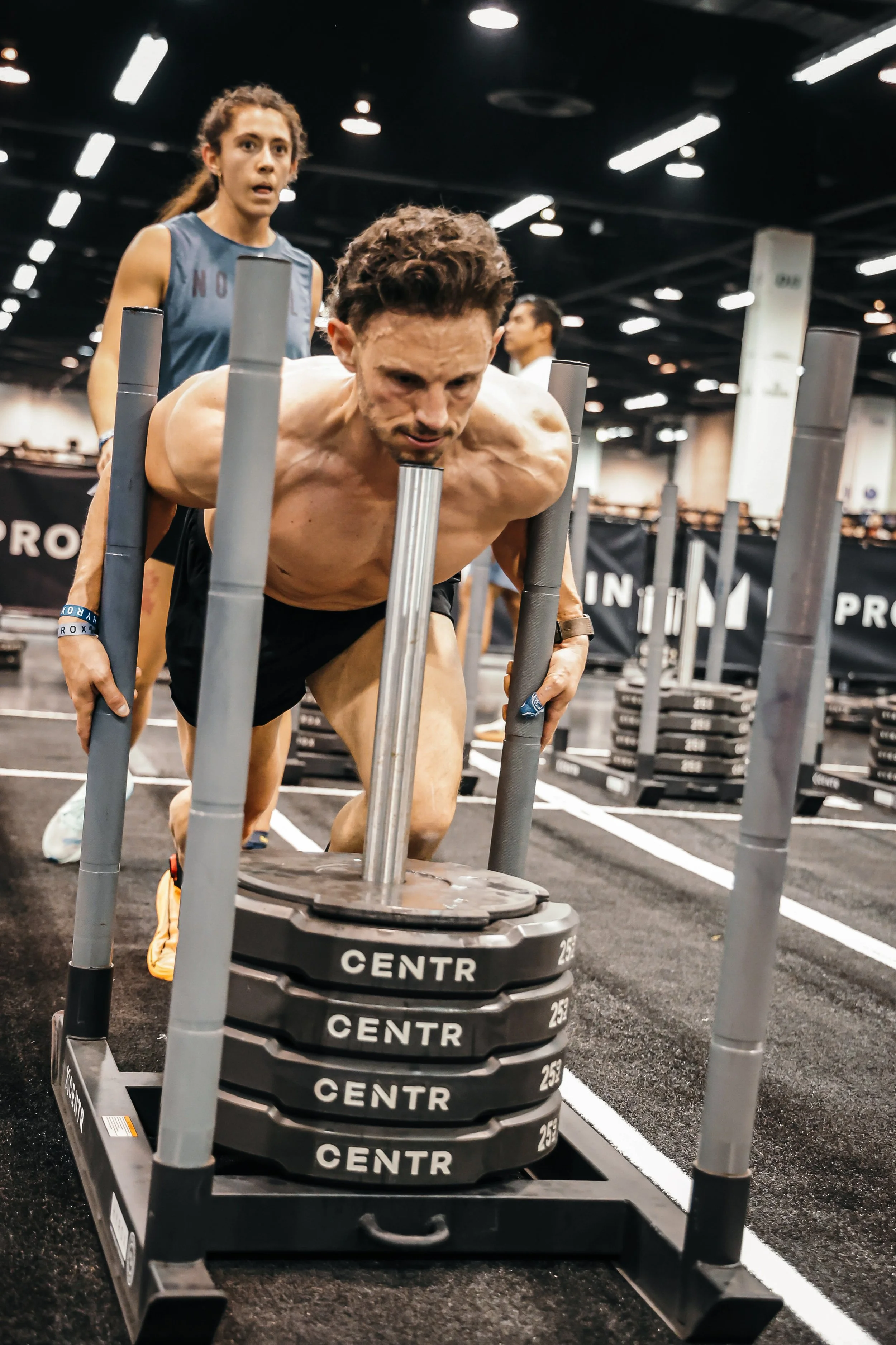 Male athlete pushing a weighted sled in a hyrox race with a doubles female partner