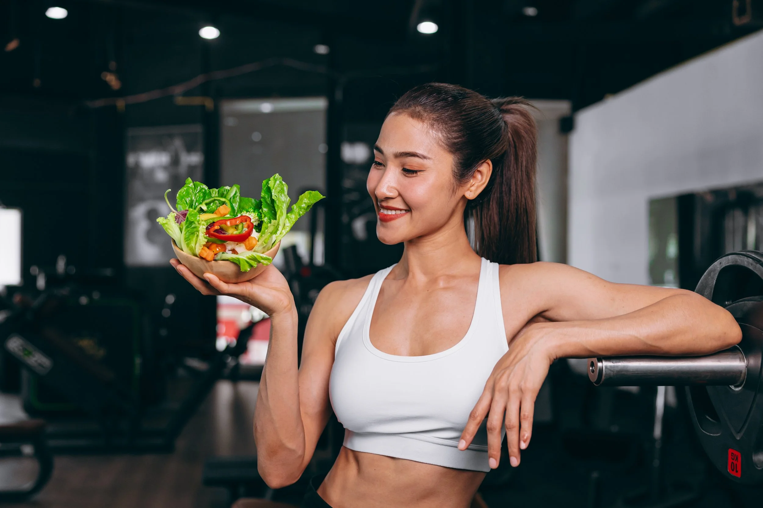 A young woman in workout attire holding a bowl of fresh vegetables and smiling at gym.