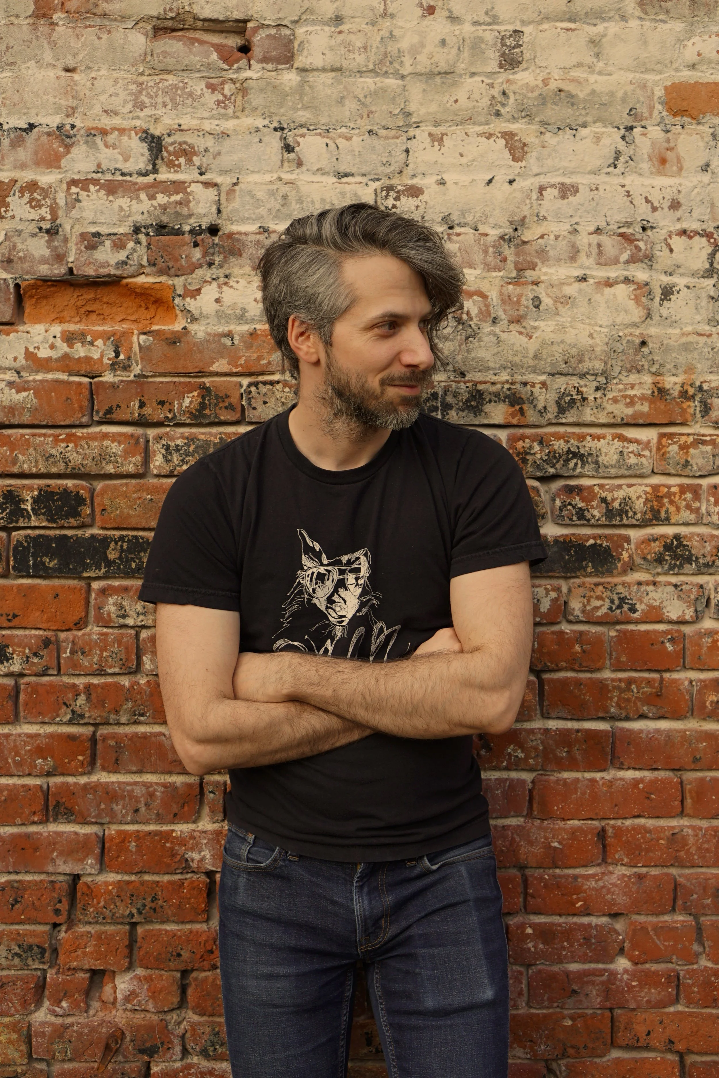 A man with gray and dark hair, beard, wearing a black graphic t-shirt and jeans, standing with arms crossed in front of an old brick wall, looking to his left.