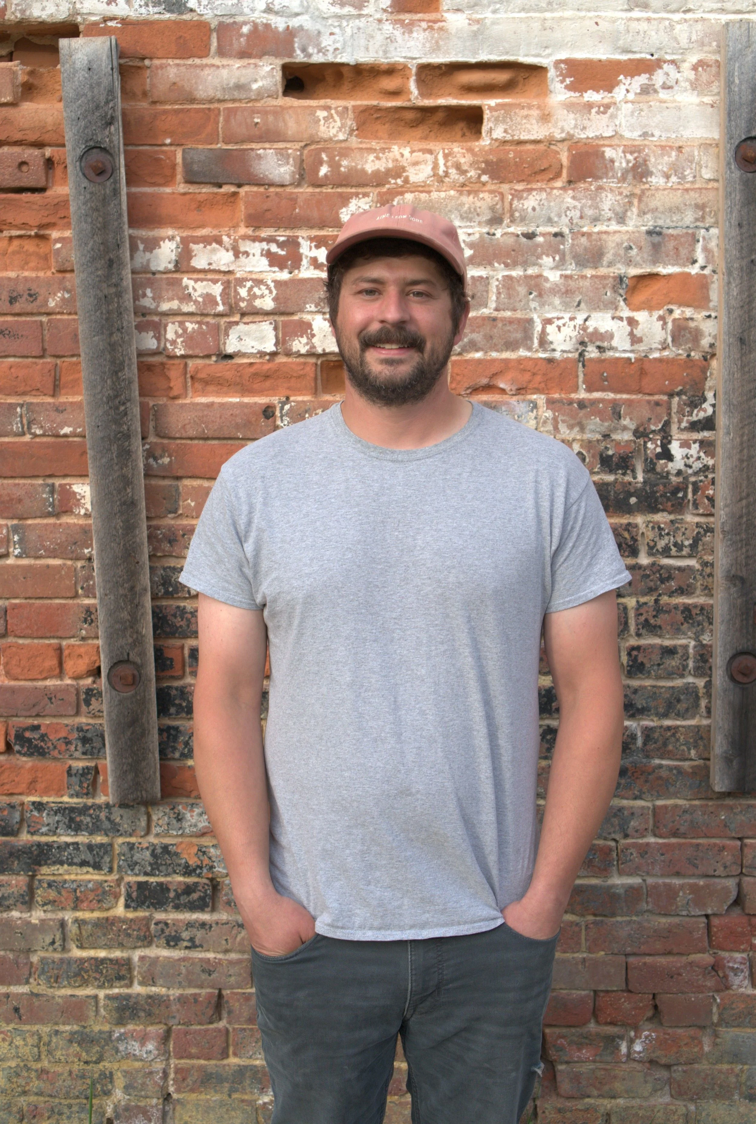 A young man with a beard and mustache wearing a gray t-shirt and a pink cap, standing outdoors in front of an old brick wall with wooden beams.