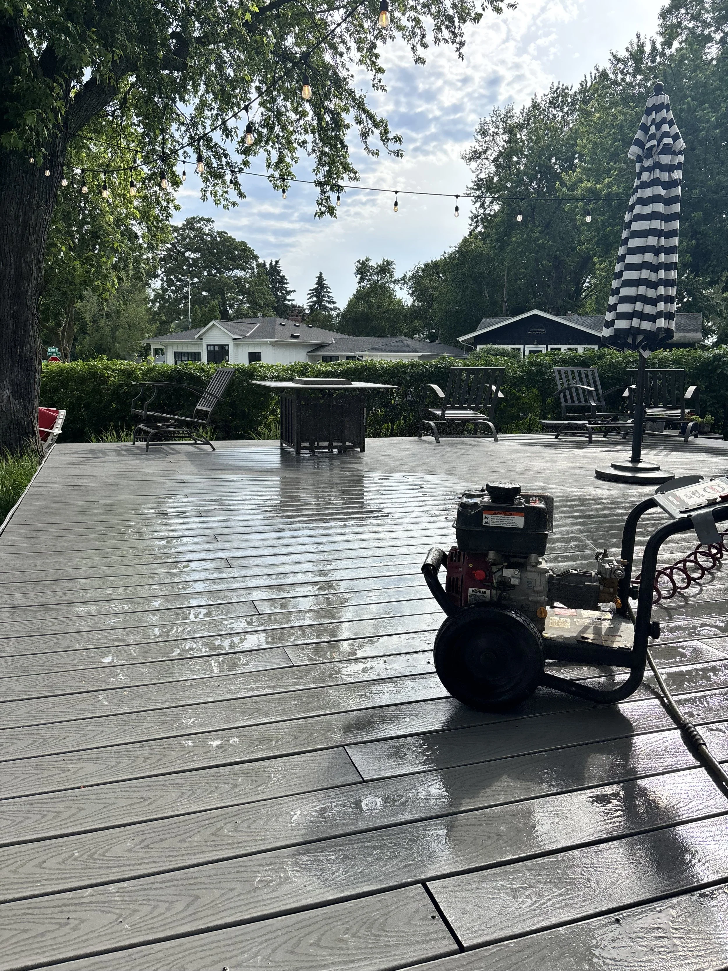 A wooden deck being washed with a power washer, with wet and dry sections visible, outdoor furniture including chairs, a table, and a striped umbrella, and string lights overhead amid trees and houses in the background.
