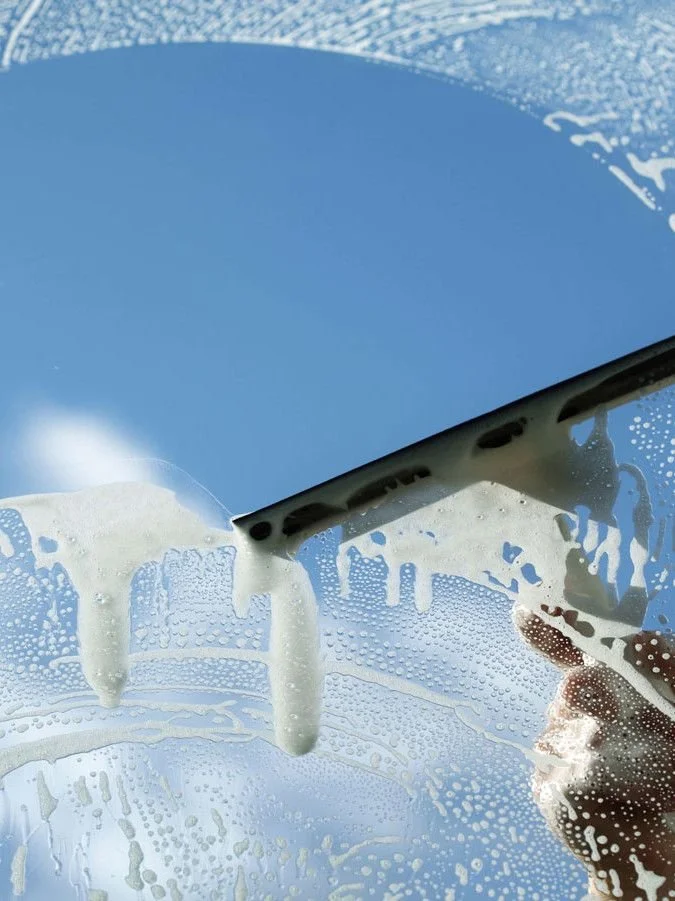 Person cleaning a car window with a squeegee, soap and water on glass, blue sky reflection