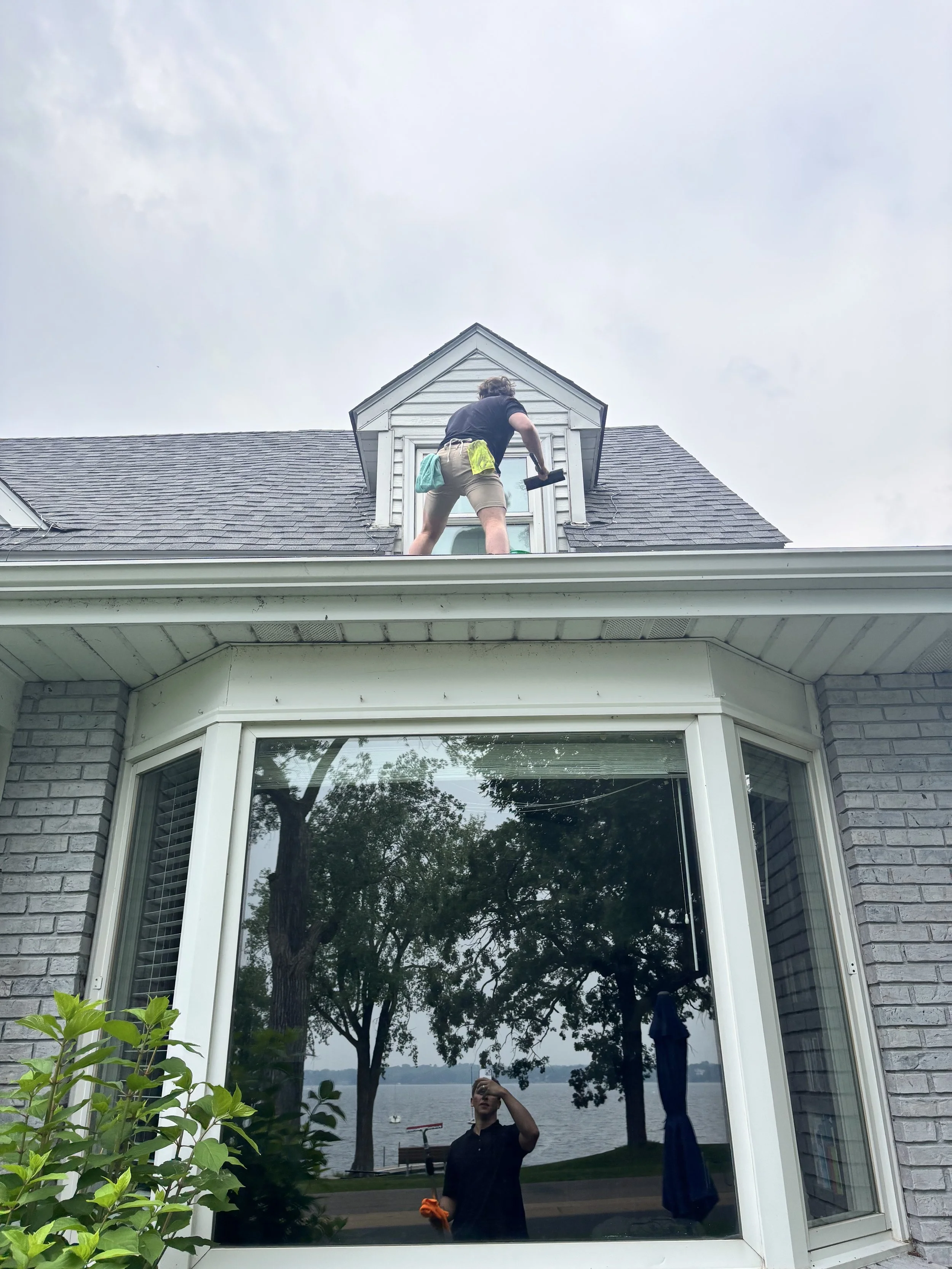 A person on the roof cleaning or repairing a window of a house, with trees and a water body visible through a large front window.