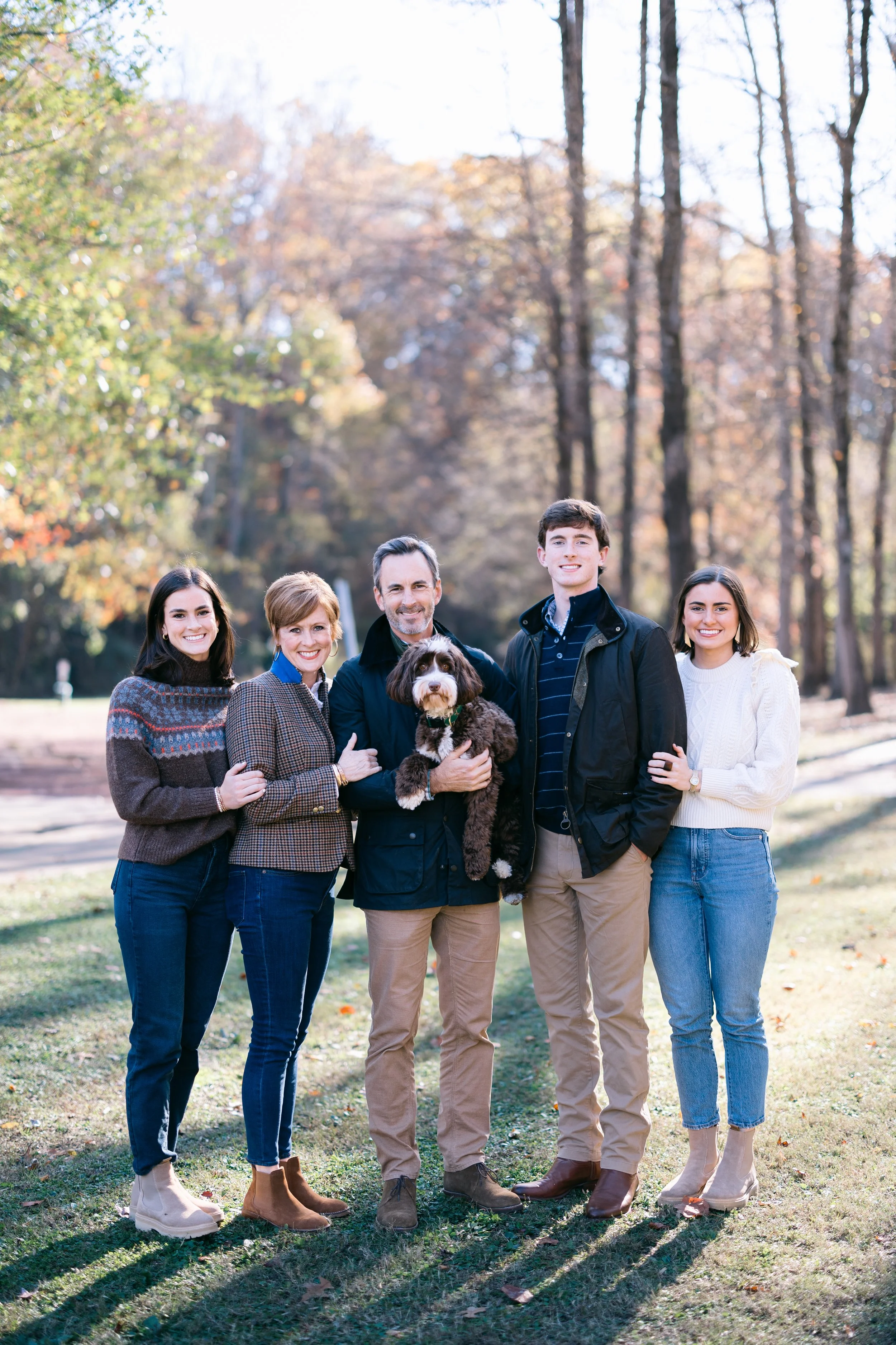 A family of five with a dog standing outdoors in a park during fall, surrounded by colorful trees.
