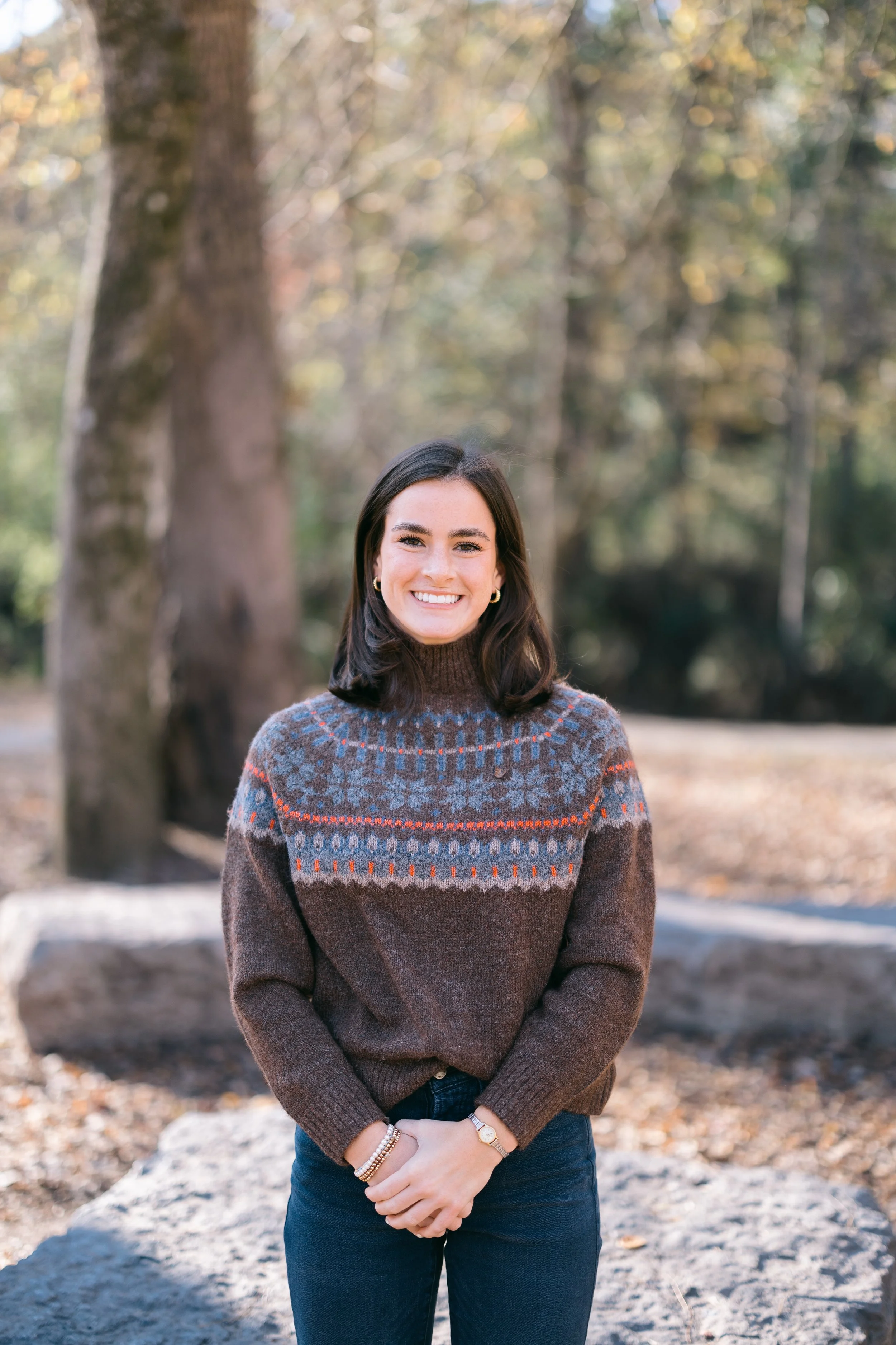 A smiling woman with dark shoulder-length hair wearing a patterned brown sweater and dark jeans stands outdoors in a park, with trees and fall foliage in the background.