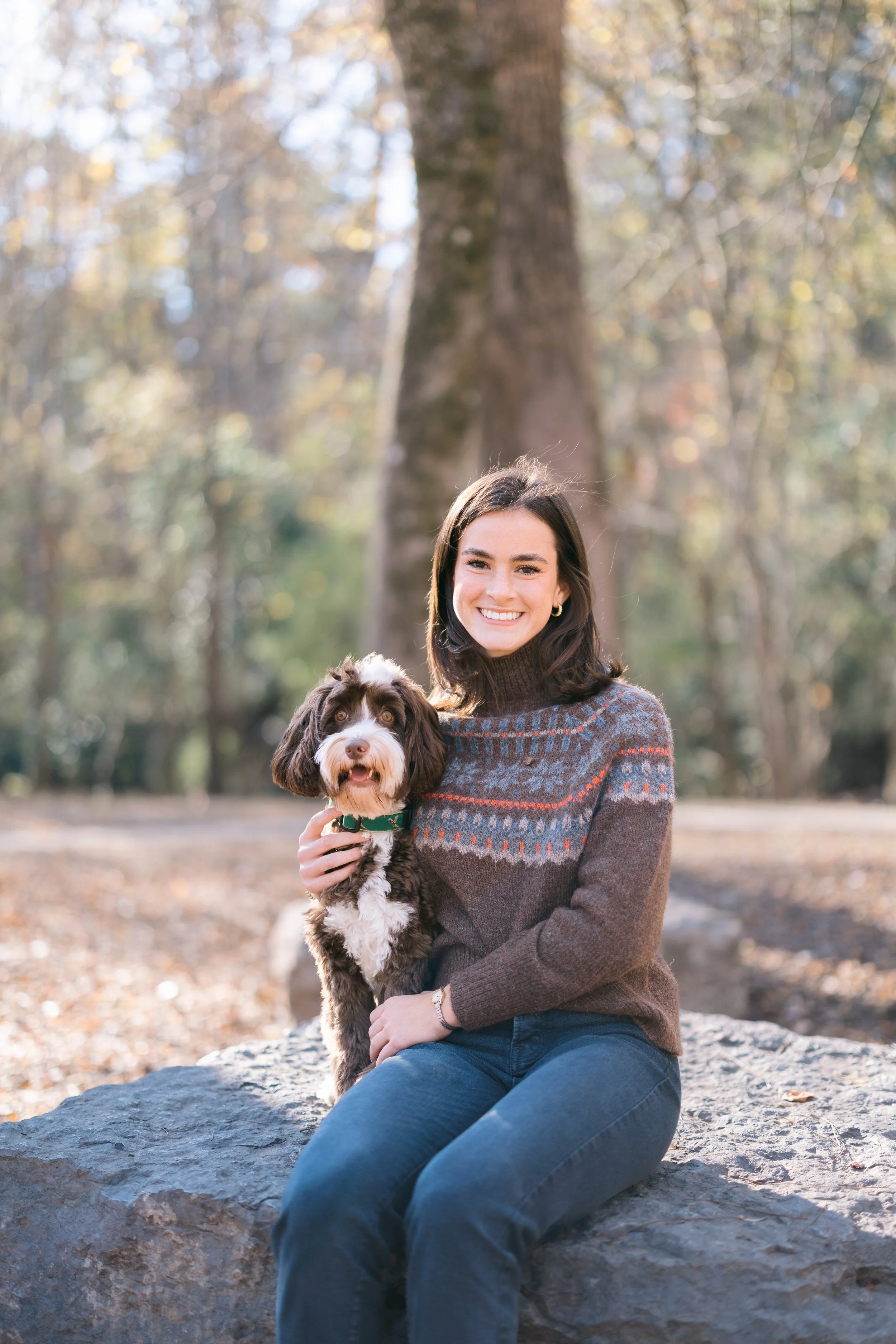 A young woman with dark hair wearing a patterned sweater and jeans sitting on a rock, holding a brown and white dog, outdoors in a wooded area with fall foliage and trees in the background.