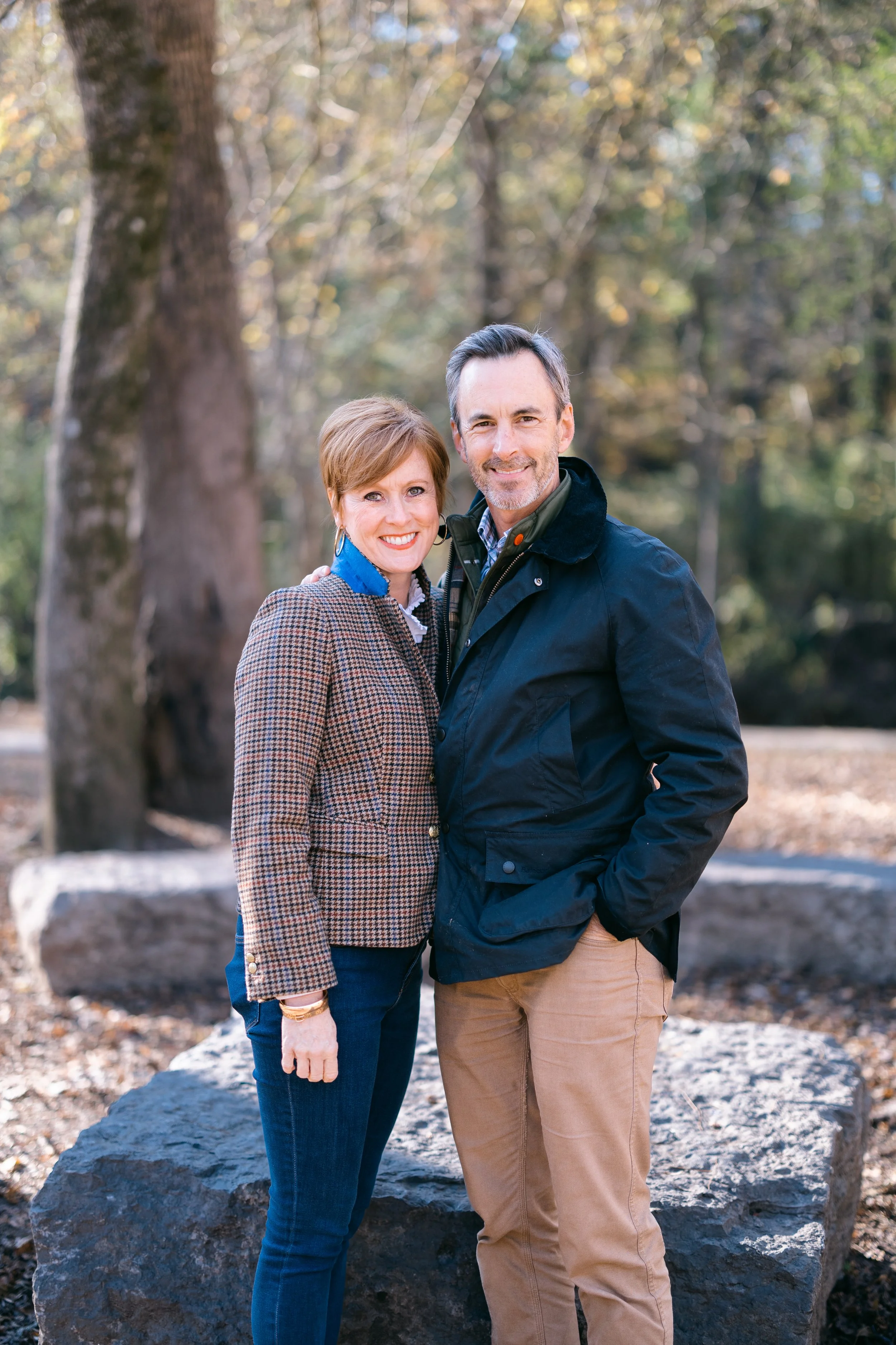A smiling middle-aged couple stands close together outdoors in a wooded area, with trees and rocks in the background, dressed in casual autumn clothing.