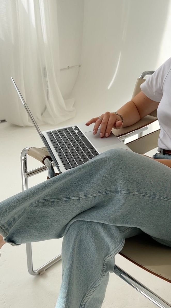A person sitting in a chair using a laptop, wearing jeans and a white shirt, in a bright room with white curtains.