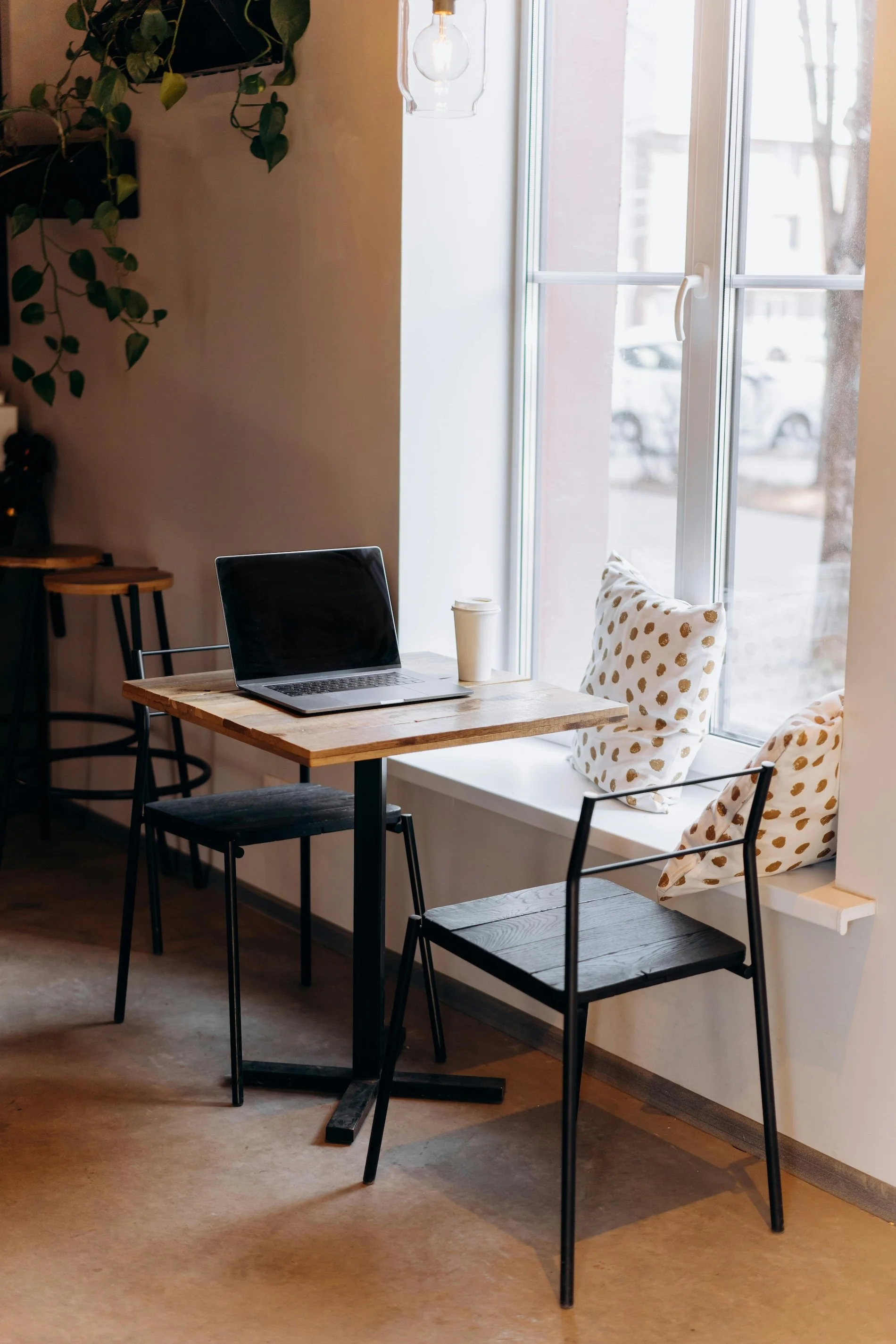 A cozy corner of a cafe with a small wooden table, two black metal chairs, a laptop, a to-go coffee cup, and decorative pillows by a large window with a soft outdoor view.