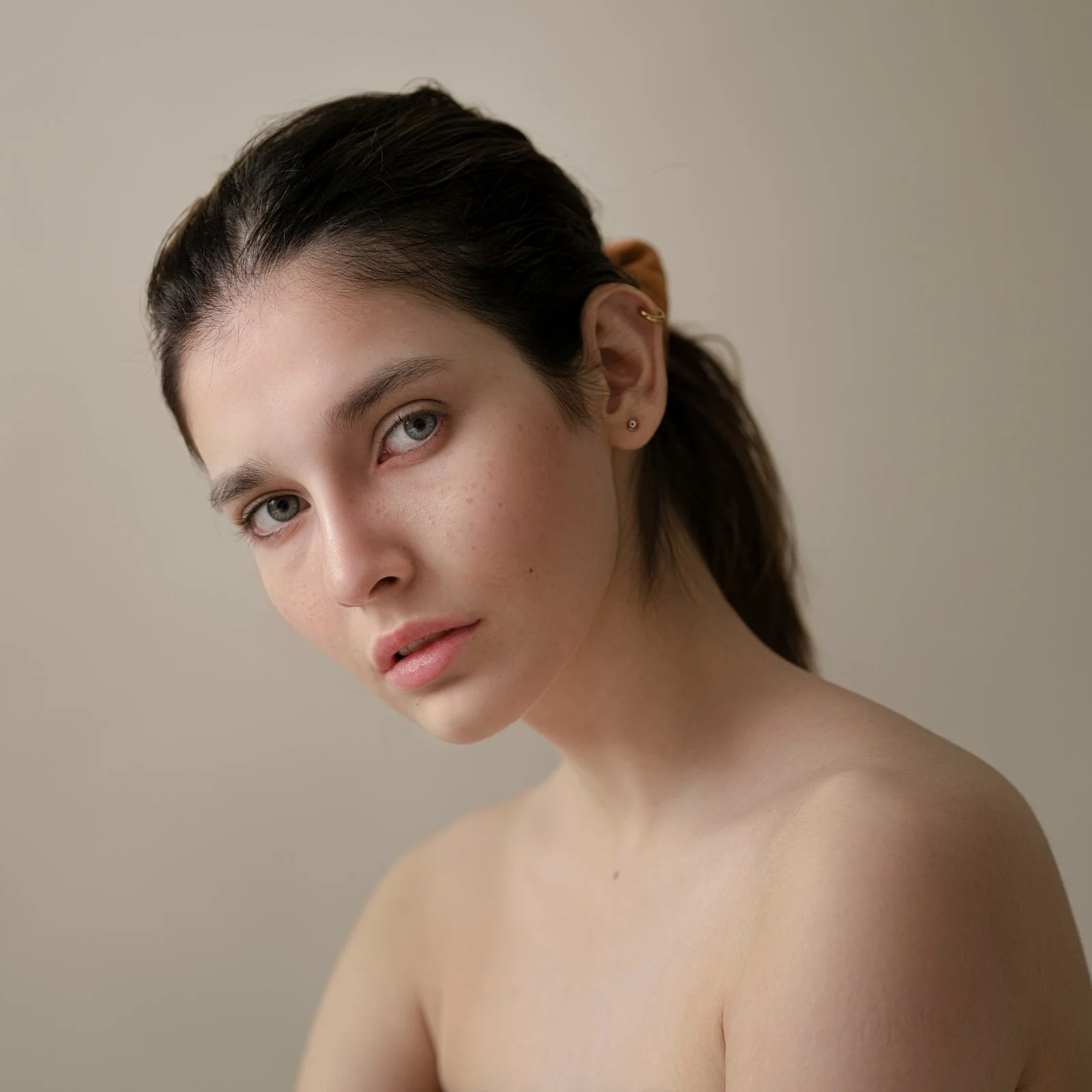 Close-up portrait of a young woman with dark hair, light skin, and blue eyes, looking at the camera with a neutral expression on a plain beige background.