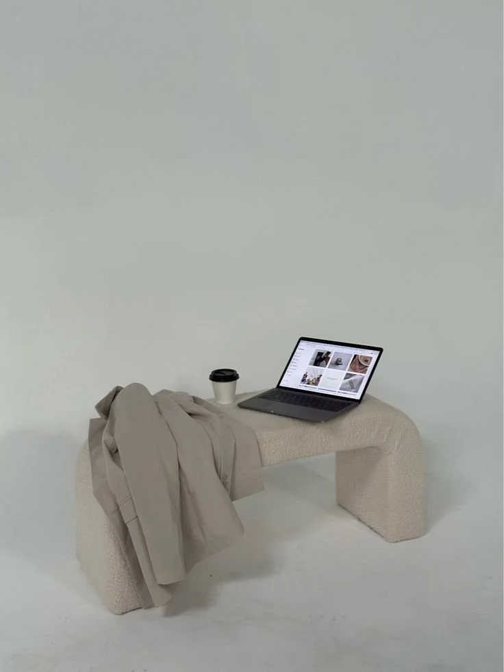 A minimalist bench with a beige coat, a coffee cup, and an open laptop on it, against a plain light background.