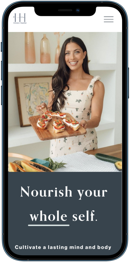 A woman with long dark hair in a floral dress holds a wooden tray with open-faced sandwiches topped with strawberries and other fruit. She is smiling in a bright kitchen with vases and framed artwork in the background. The mobile screen shows a website with the slogan "Nourish your whole self" and a subtitle about cultivating a lasting mind and body.