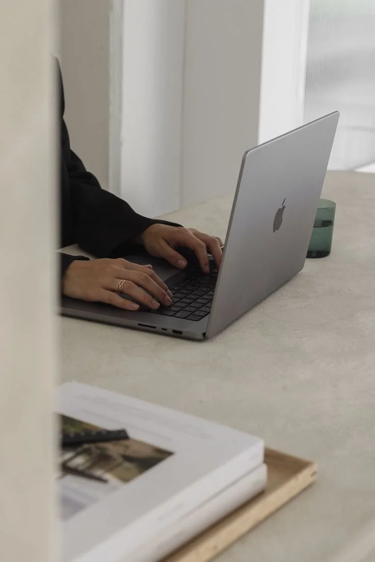 Person typing on a silver MacBook laptop at a white table with a green mug and a stack of papers nearby.
