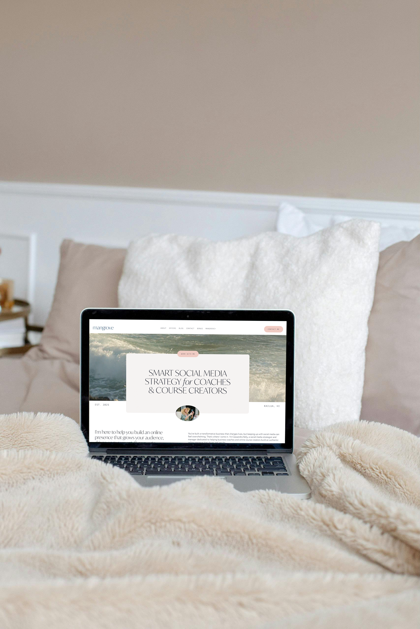 Laptop resting on a cozy bed with beige and white pillows, showing a website titled 'SMART SOCIAL MEDIA STRATEGY for COACHES & COURSE CREATORS' with a beach background.