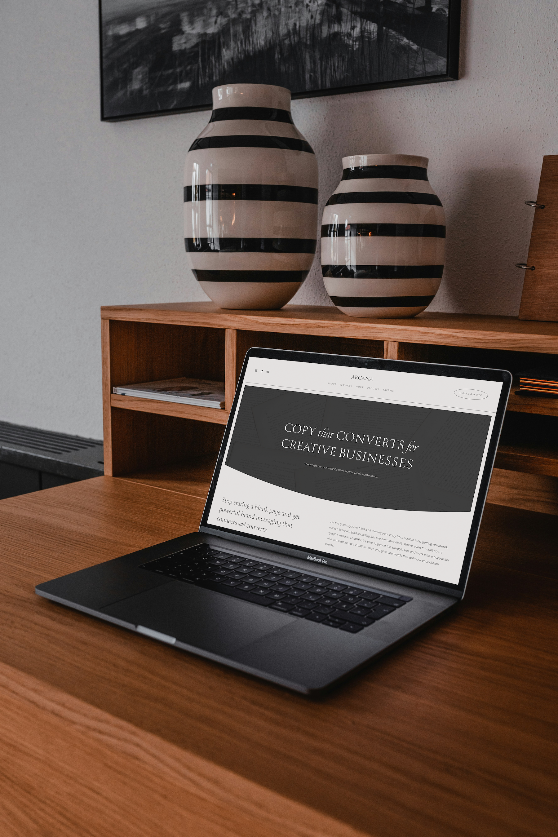 A MacBook Pro laptop open on a wooden desk displaying a website titled 'COPY that CONVERTS for CREATIVE BUSINESSES.' Behind the laptop, there is a wooden cubby shelf with a few books or papers inside. On top of the shelf, there are two large, matching ceramic vases with black and white horizontal stripes. A black and white framed aerial photograph or artwork hangs on the wall above the vases.