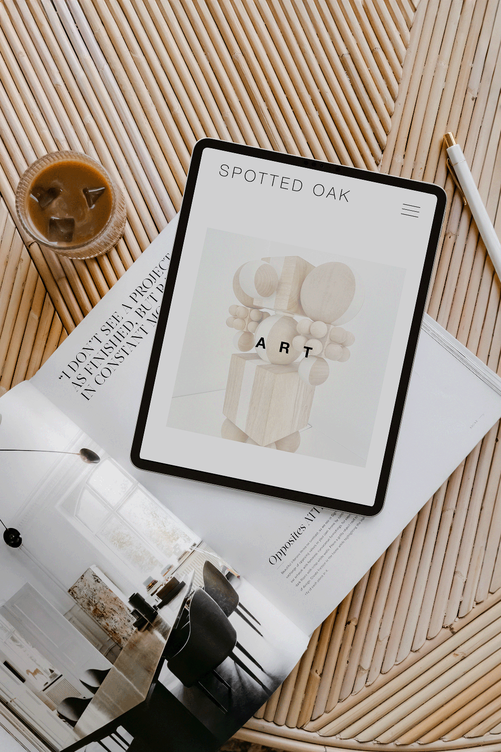 An iPad displaying an art website with a wooden sculpture, a glass of iced coffee on a wooden table, a white pen, and an open magazine with interior design photos.