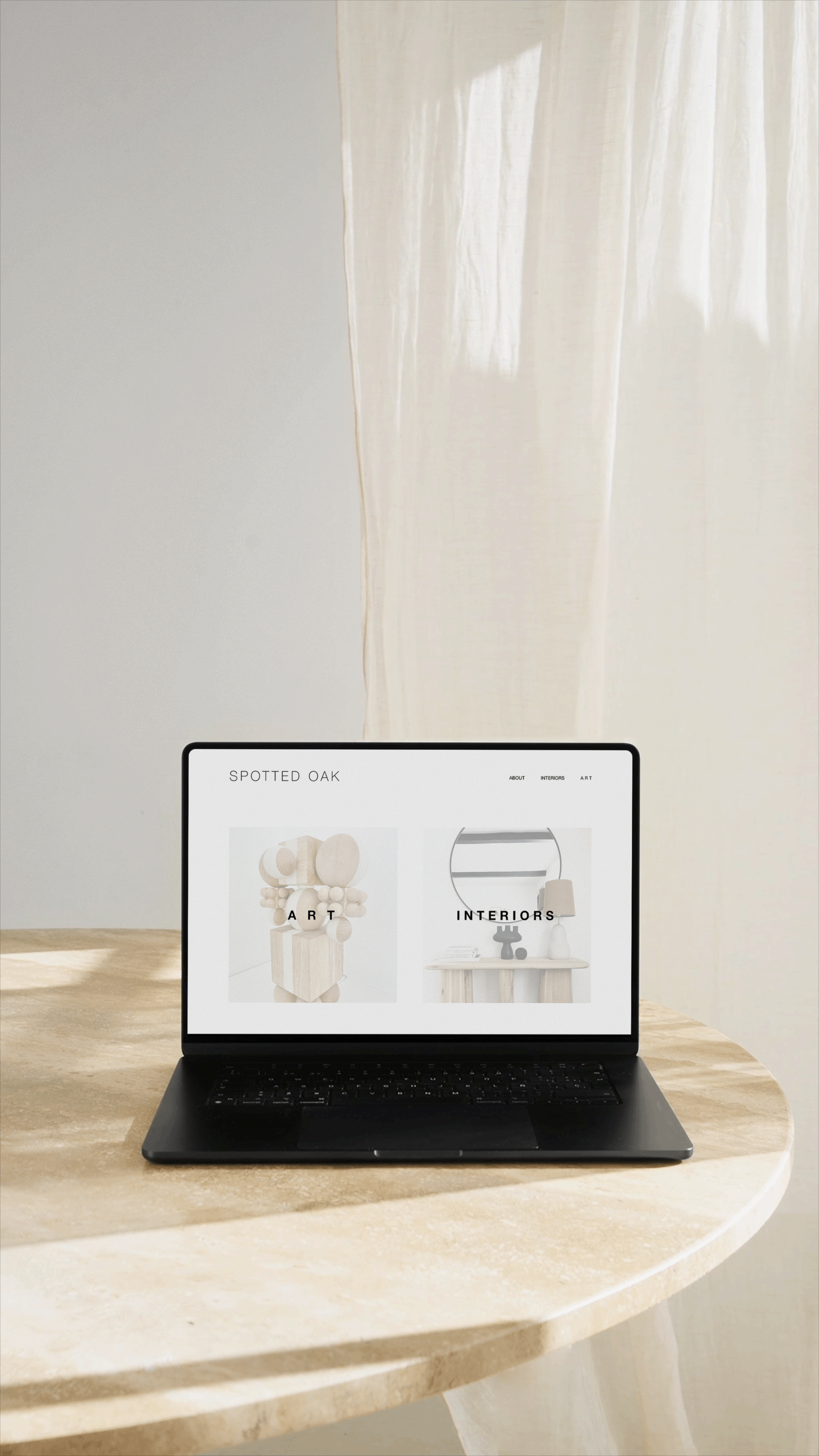 A black laptop on a light-colored, rounded wooden table displaying a website for Spotted Oak, with sections labeled Art and Interiors. A beige curtain and a light-colored wall are in the background.