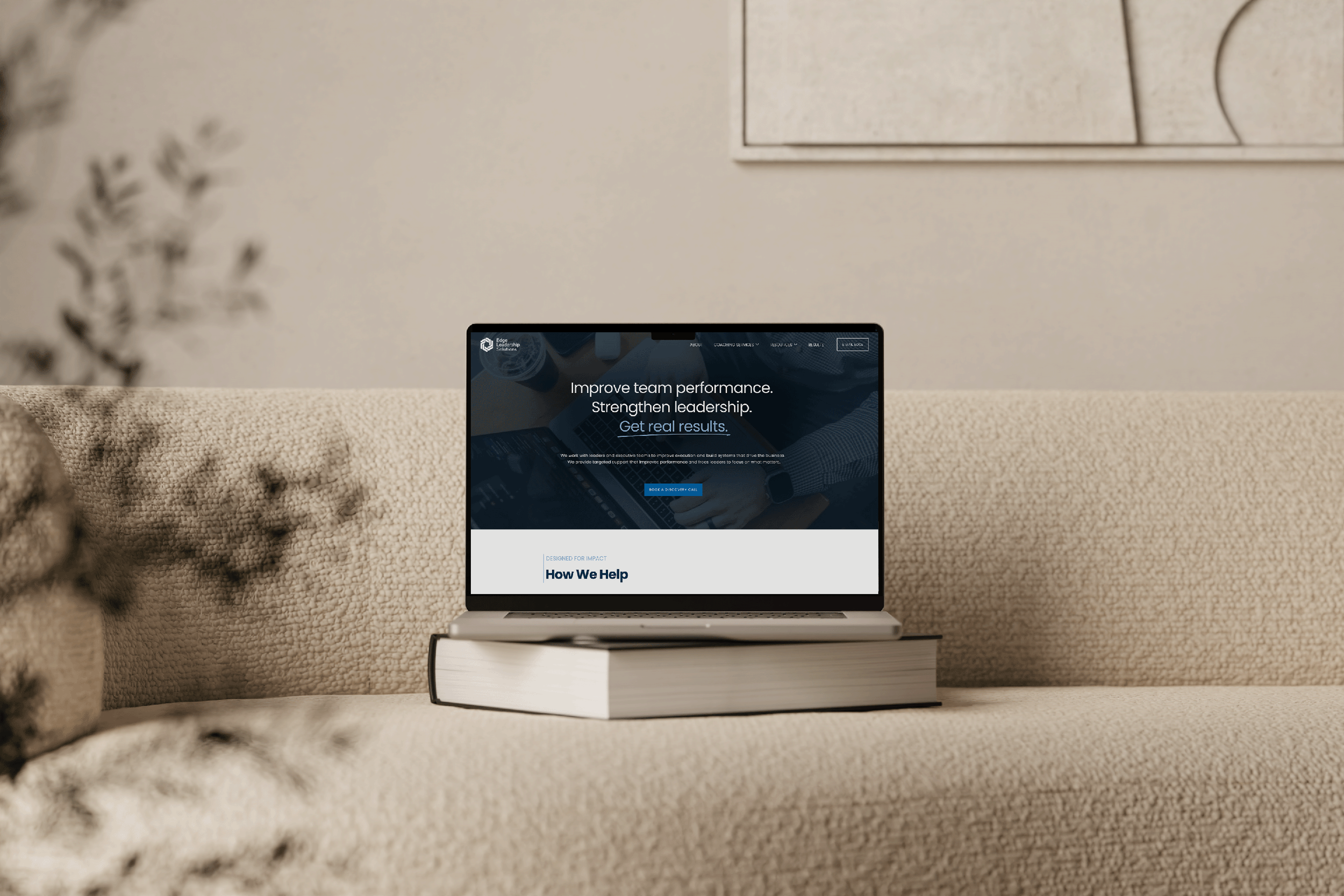 Laptop on a closed book placed on a beige fabric sofa, displaying a website about improving team performance and leadership.