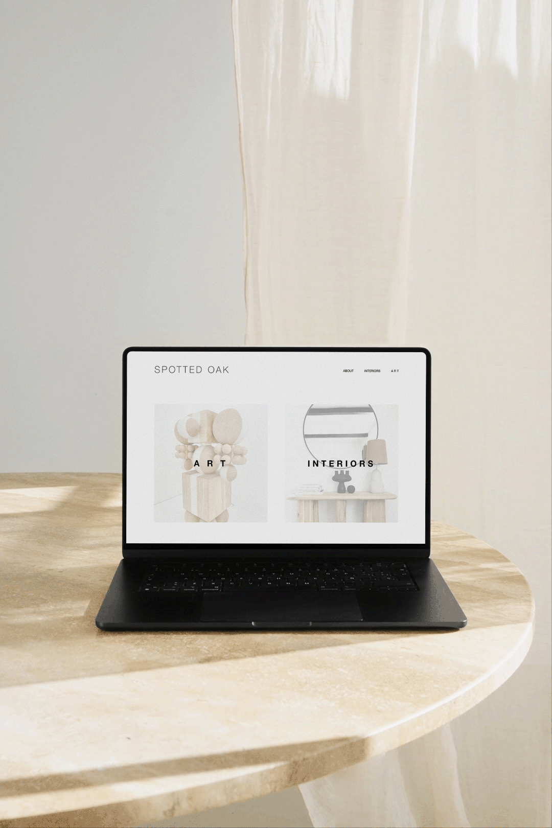 A laptop on a round, beige, marble table displaying a website titled 'Spotted Oak' with sections for art and interiors, next to sheer, cream-colored curtains.