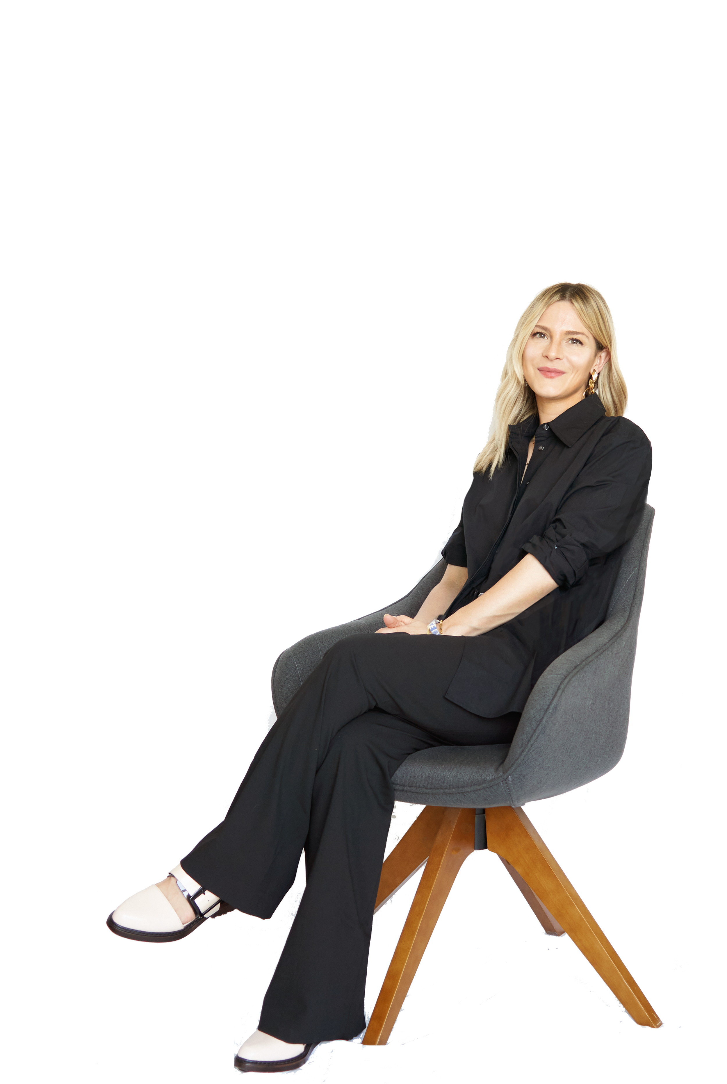 A woman with blonde hair sitting in a grey armchair, wearing a black button-up shirt, black pants, and white shoes with black accents, smiling at the camera.