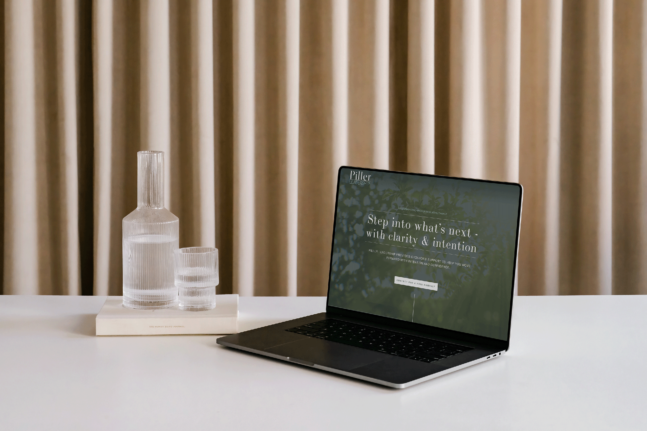 A laptop on a white table with a green website displayed on the screen. Next to the laptop, there are three transparent glass bottles of different sizes on a white book.